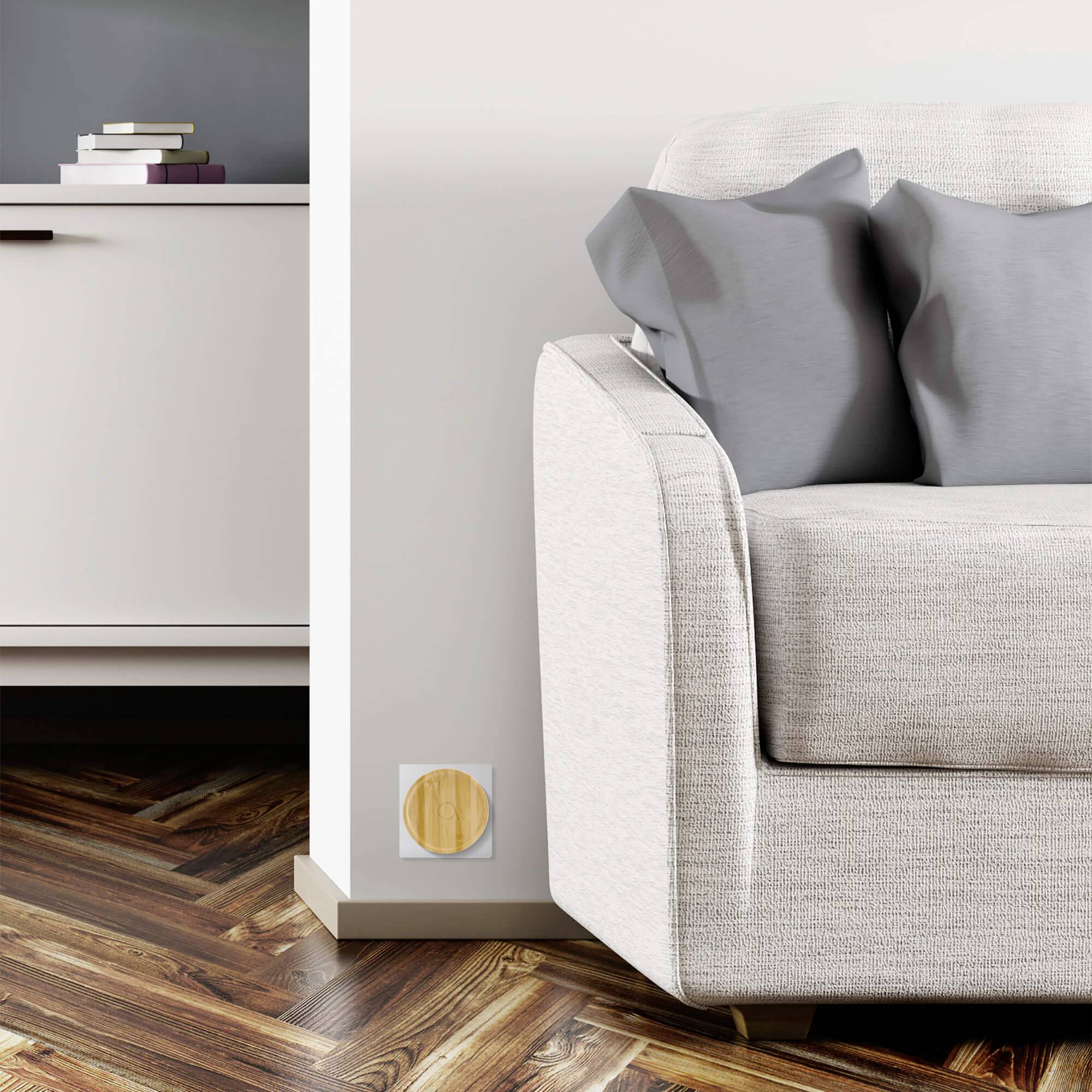 Een grijze bank met twee kussens staat op een houten visgraatvloer naast een witte muur. Een klein houten rek is zichtbaar.