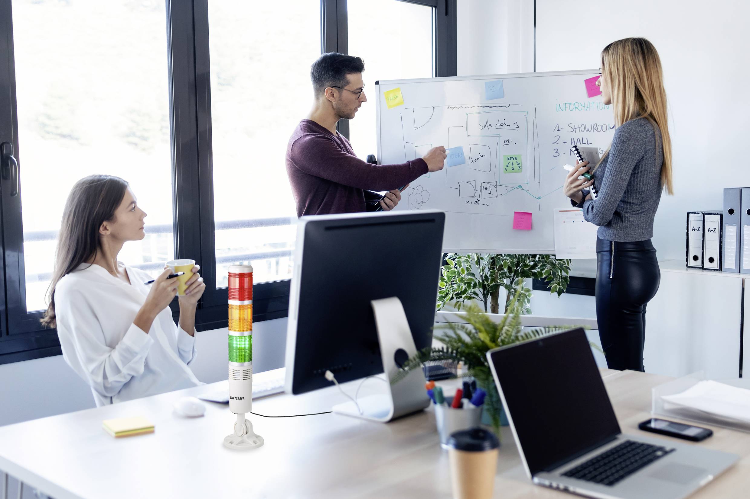 'Drie personen in een kantoor: Twee staan bij het whiteboard, bespreken grafieken en aantekeningen; een zit aan tafel met een kopje koffie.'