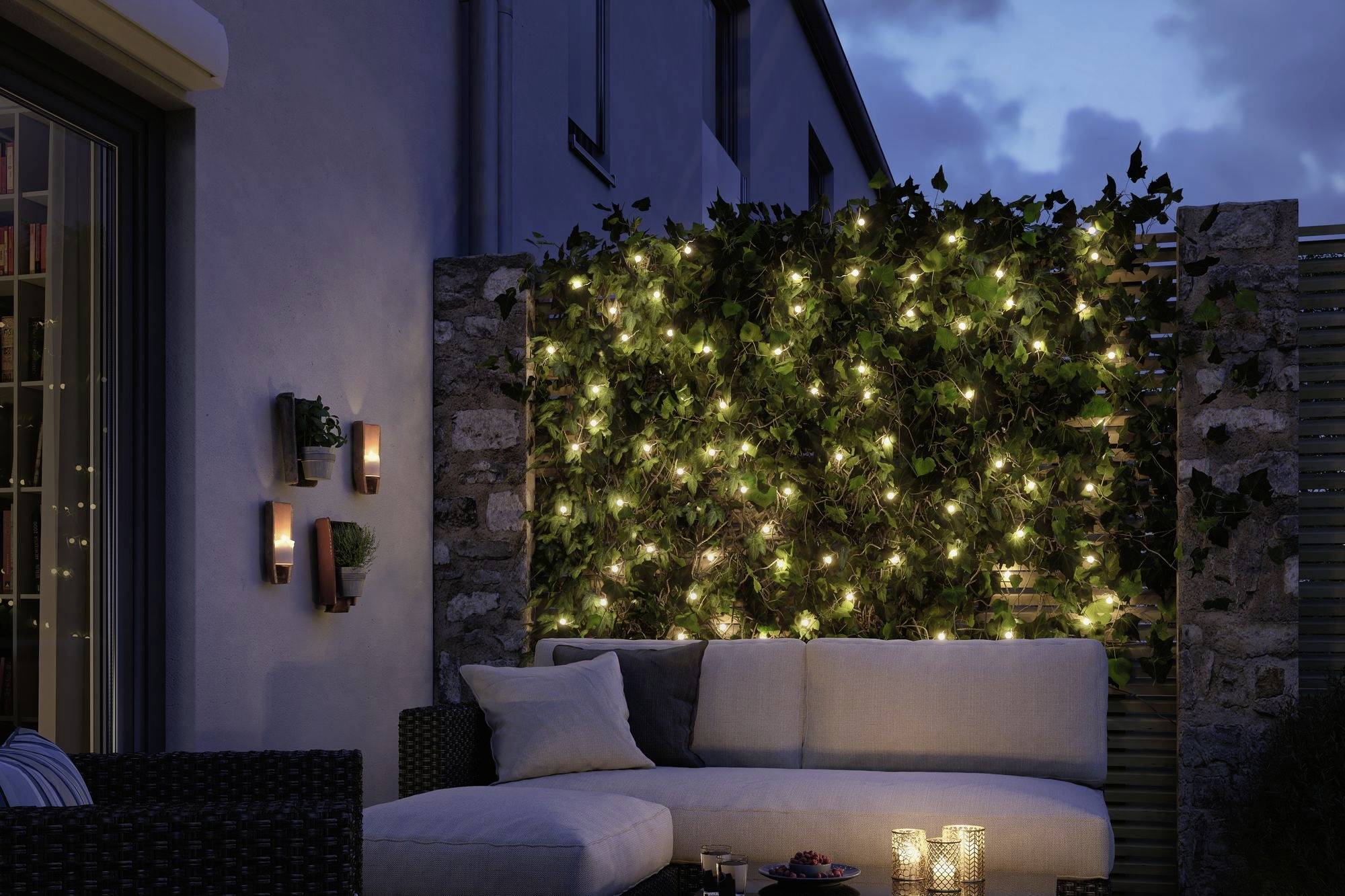Gezellig terras bij avondschemering, met slingers van lichtjes en kussens op de bank. Kaarsen in glazen vervolmaken de sfeervolle verlichting.