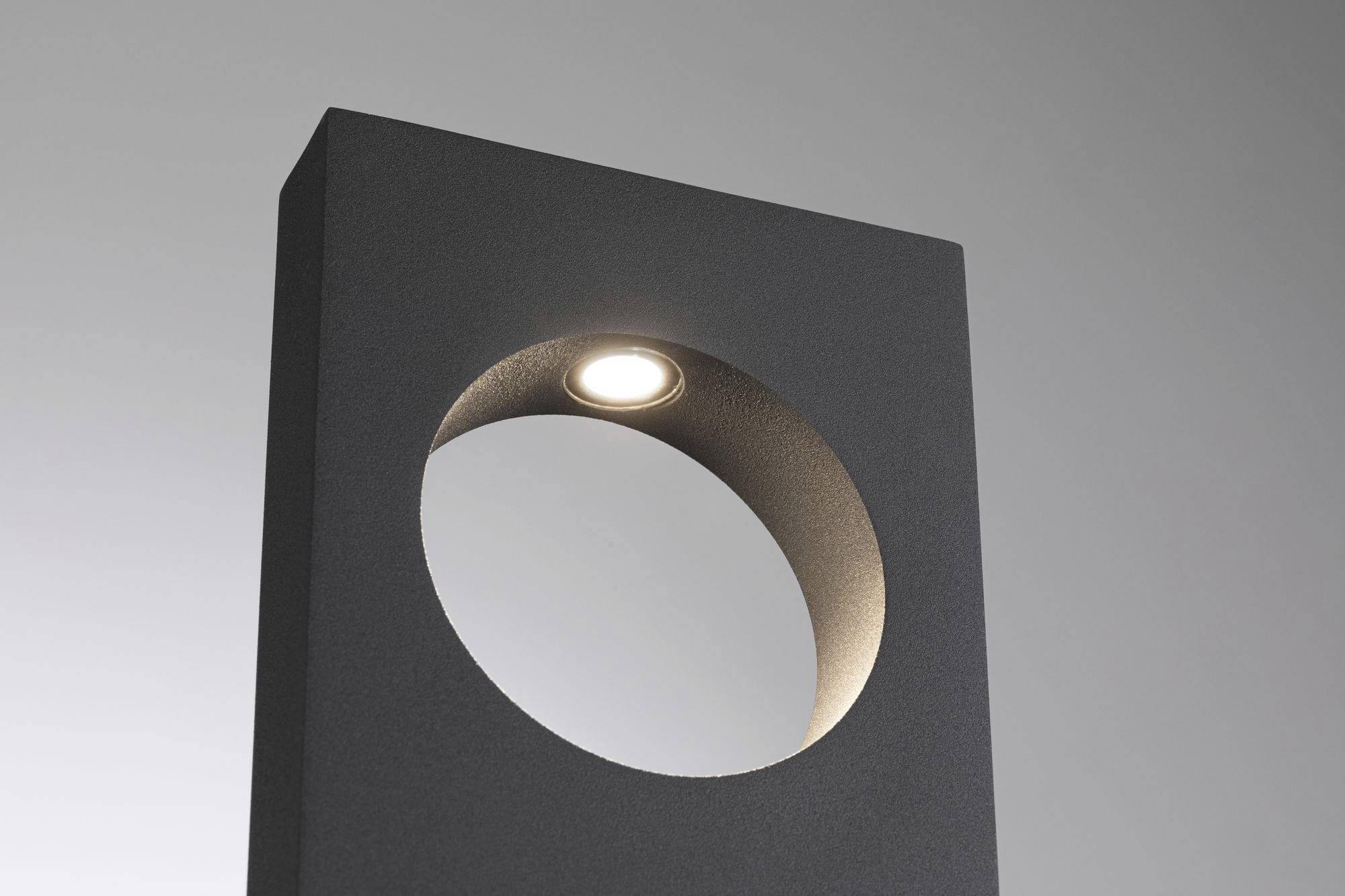 Een rechthoekig, donker object met een rond uitgesneden deel in het midden, bovenaan een ingebouwde verlichting. De achtergrond is lichtgrijs.