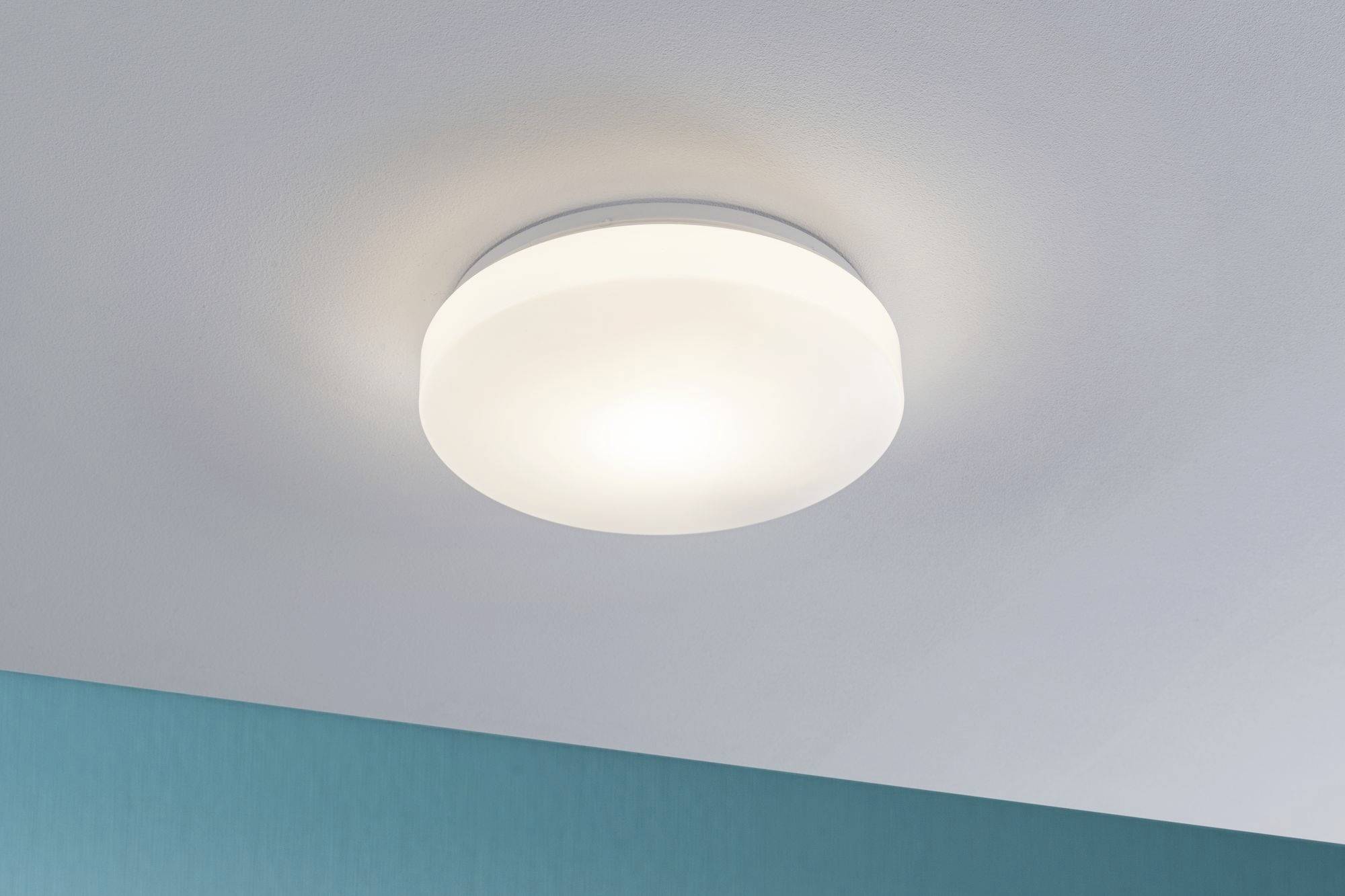 Ronde plafondlamp, verlicht, aan een wit geschilderd plafond boven een turquoise muur.