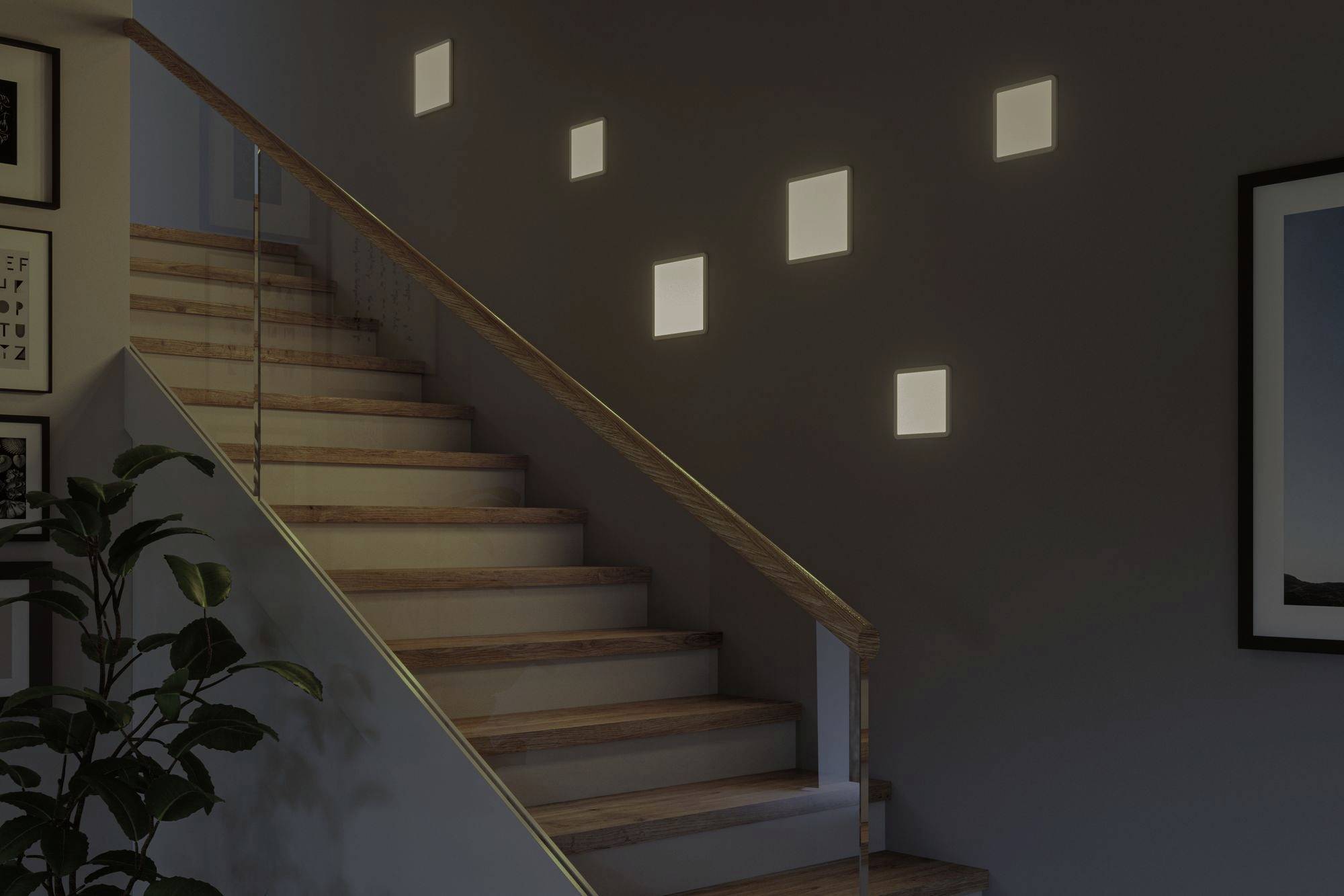 Een modern trappenhuis met verlichte vierkante lampen aan de muur, planten en ingelijste schilderijen op de achtergrond.
