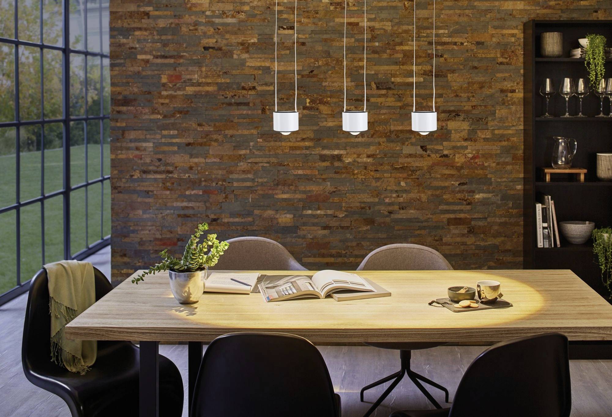 Eetkamer met houten tafel, vijf stoelen en drie hangende lampen. Openliggend boek en planten op tafel, bakstenen muur op de achtergrond.