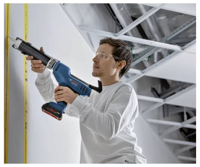 Een persoon in veiligheidsuitrusting gebruikt een elektrisch gereedschap om materiaal aan te brengen langs een hoeknaad in een bouwkundige omgeving. Plafondconstructie zichtbaar boven de werkplek.