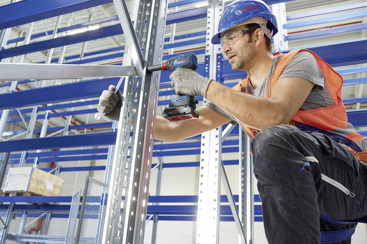 Arbeider met veiligheidshelm en vest monteert een metalen rek met een accuschroevendraaier in een magazijn, symboliseert efficiëntie en veiligheid.