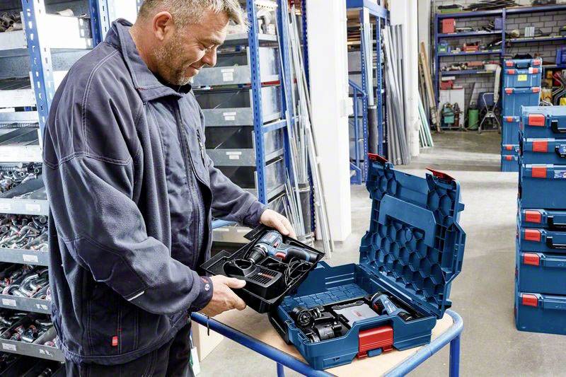Een man in werkkleding controleert boren in een open gereedschapskoffer in een opslagruimte met schappen vol andere gereedschapskoffers.
