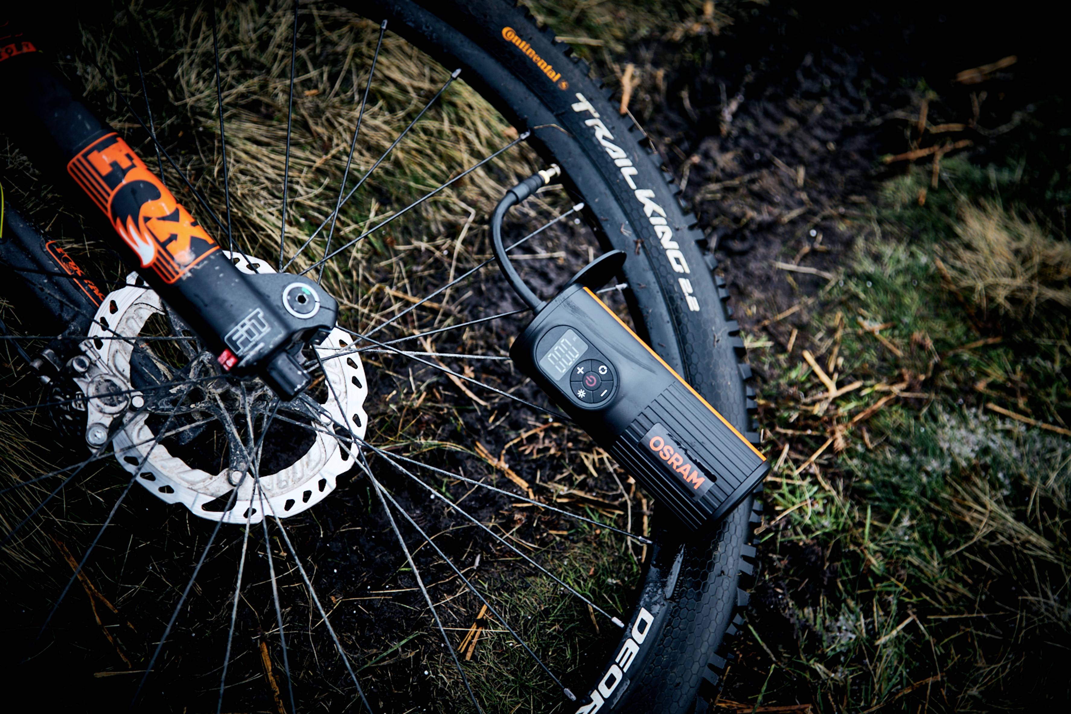 Fietsband opgepompt met een zwarte elektrische luchtpomp op modderige grond buiten. Links is de voorvork zichtbaar.