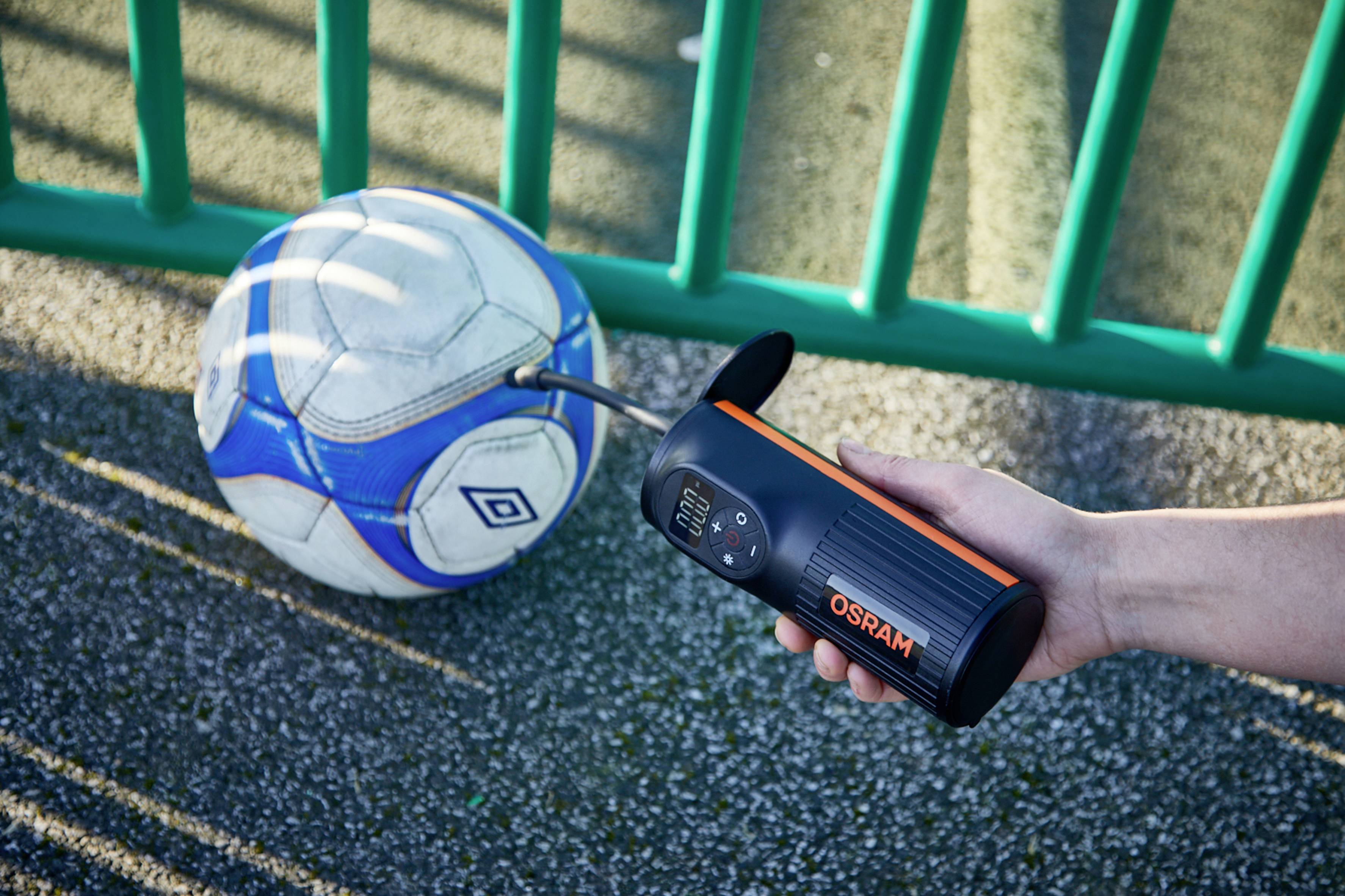 Een persoon pompt een voetbal op met een draagbaar apparaat, terwijl de bal op een bestrate oppervlakte naast een groene hek ligt.