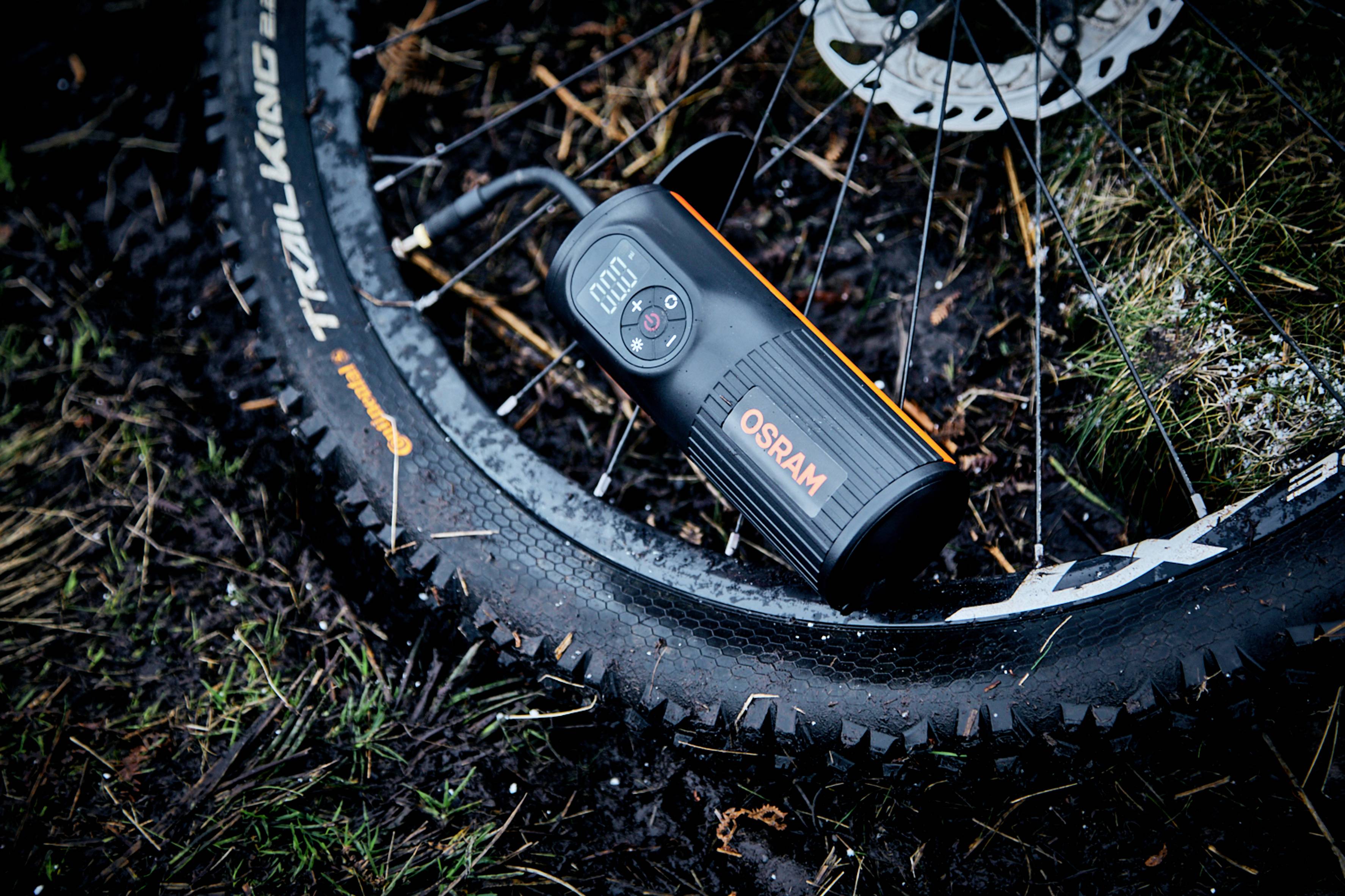 Een draagbare compressor pompt een fietsband op. Op het display staat '0.00'. Omgeving is vochtige bosbodem.