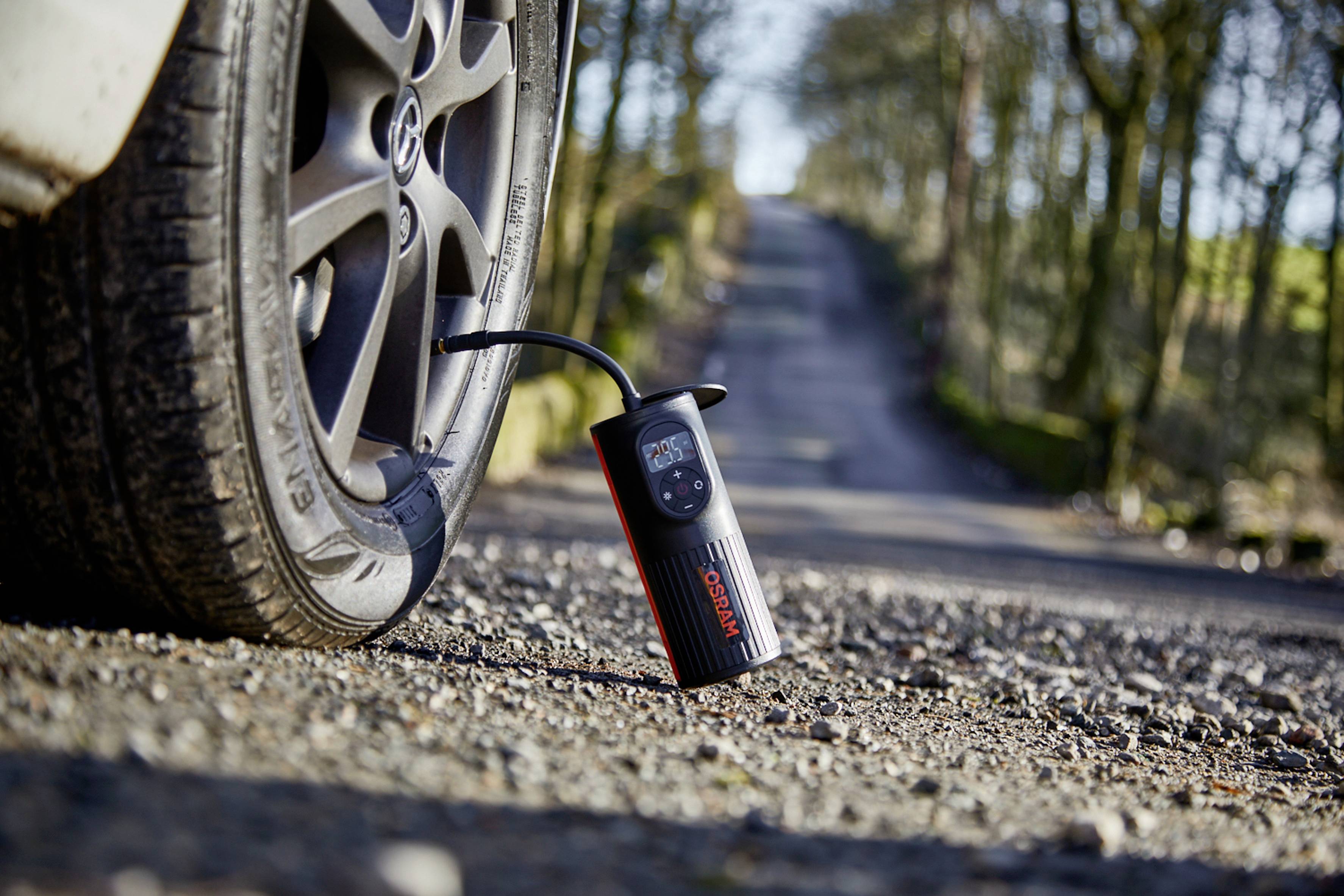 Een digitale luchtcompressor vult een autoband langs een landelijke weg. Omgeving met bomen op de achtergrond.