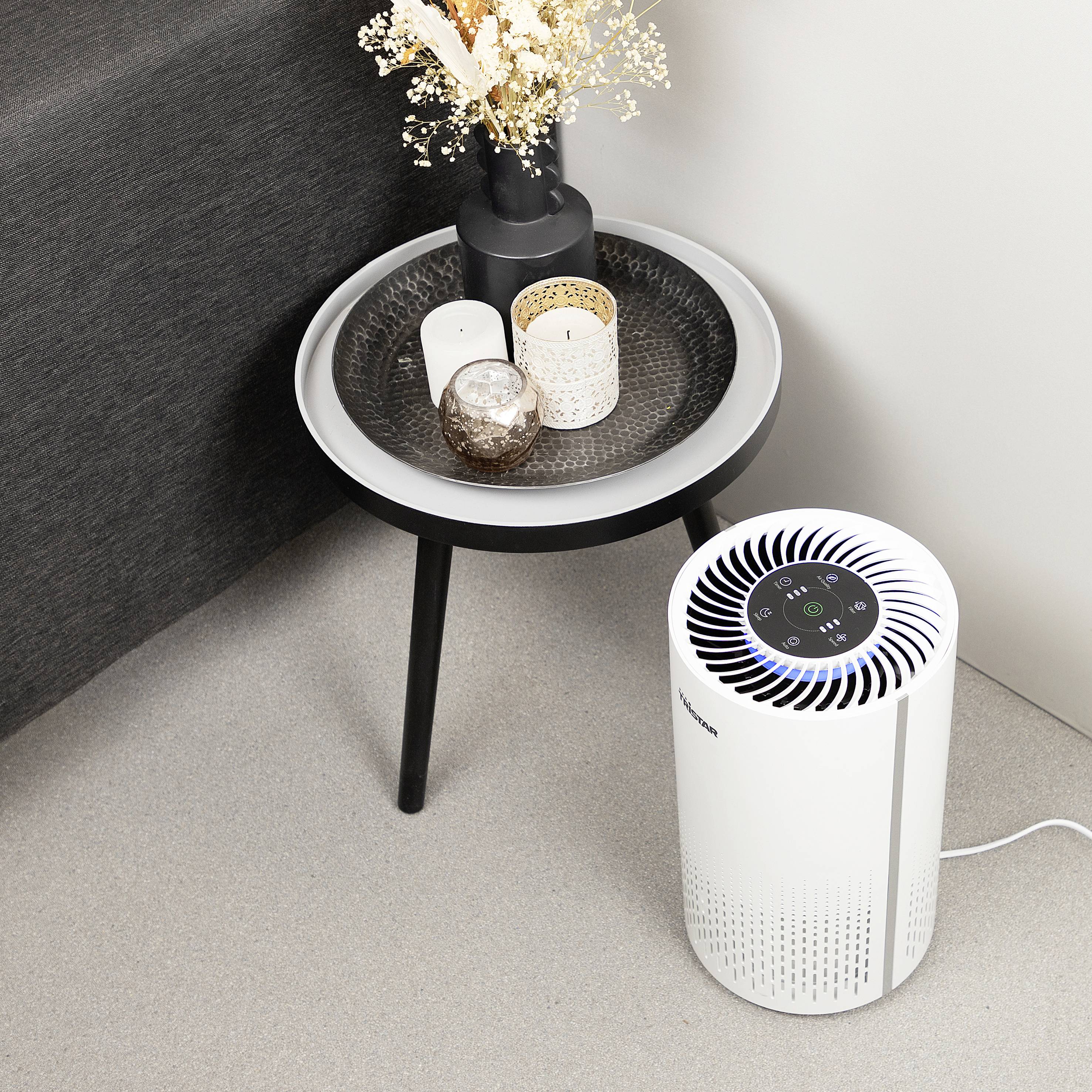 Een luchtzuiveraar staat op de vloer naast een klein bijzettafel met kaarsen en gedroogde bloemen in een vaas.