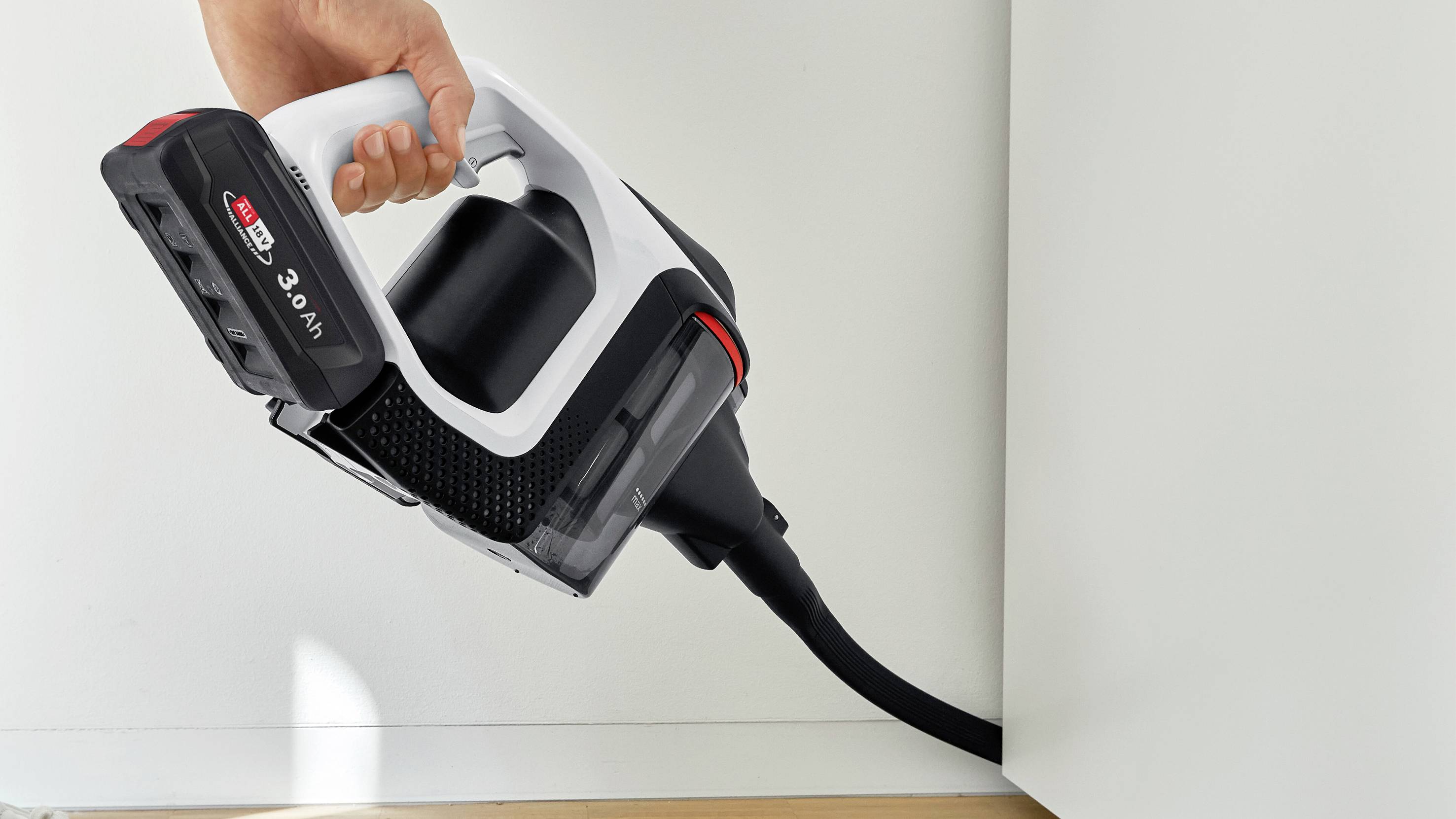 Een hand bedient een draadloze stofzuiger die een hoek tussen muur en vloer schoonmaakt.