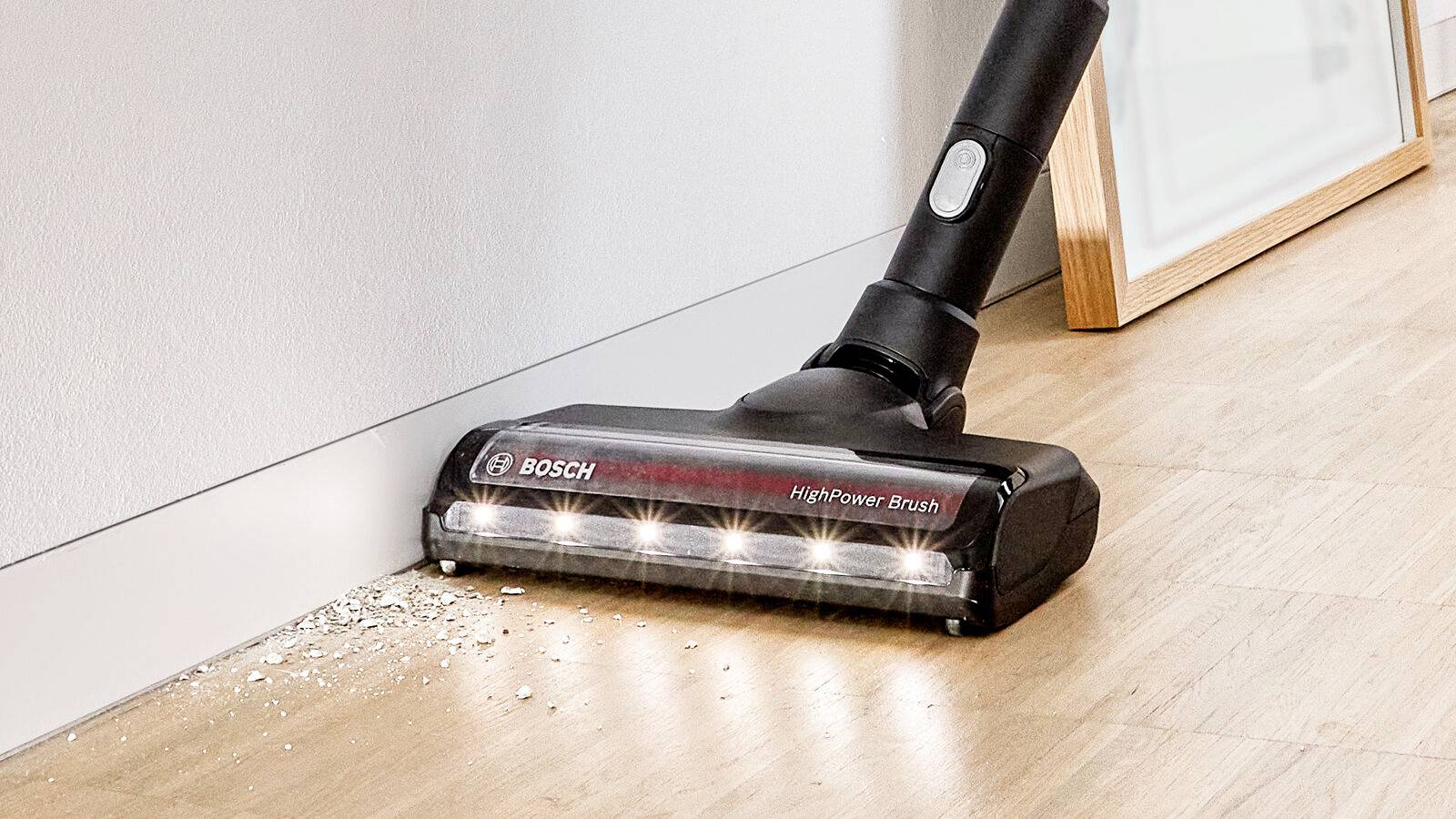 Een stofzuiger met LED-verlichting verwijdert kruimels op een houten vloer langs een muur.