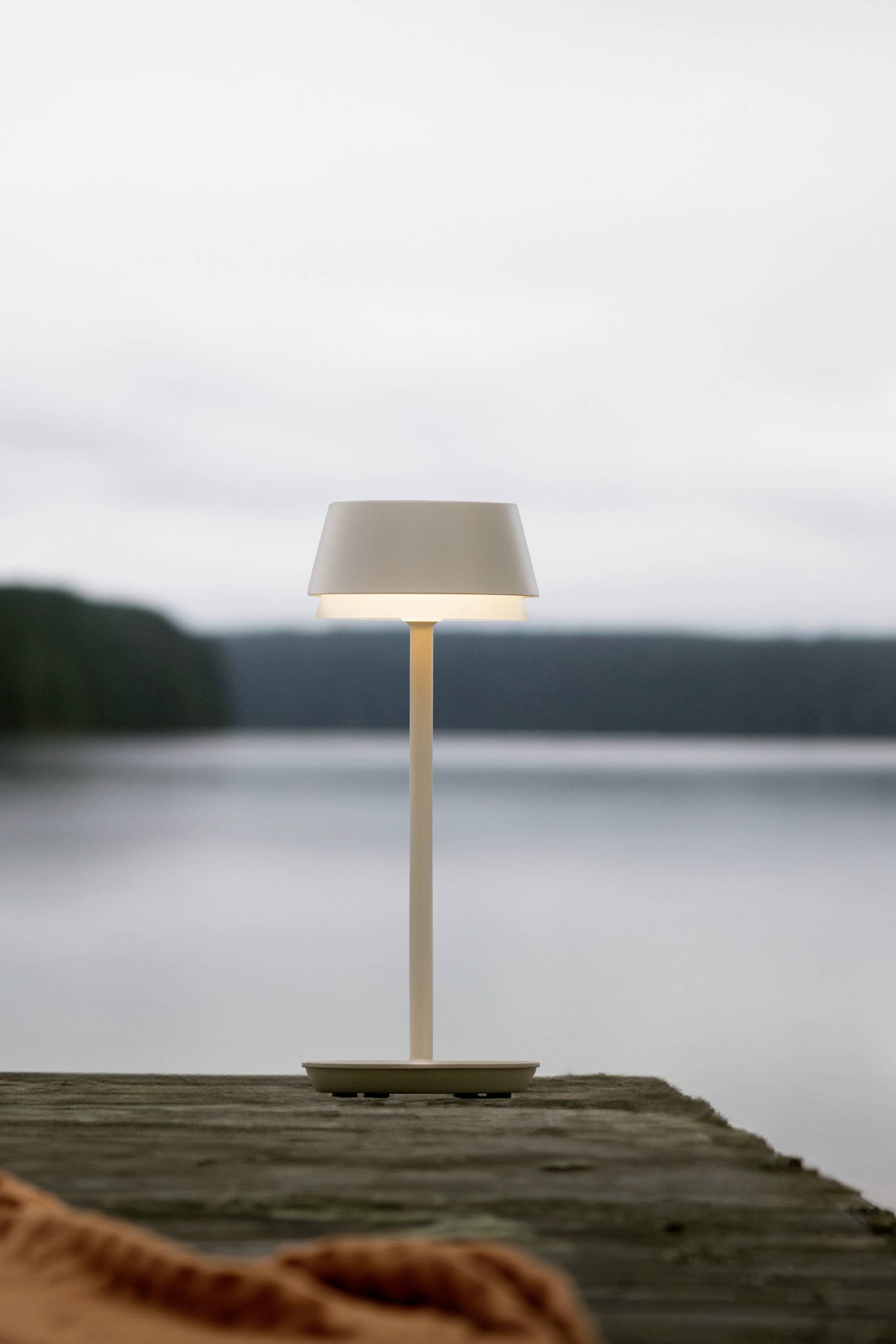 Een slanke, moderne tafellamp schijnt op een houten terras aan de rustige oever van het meer. Op de achtergrond zijn beboste heuvels zichtbaar.