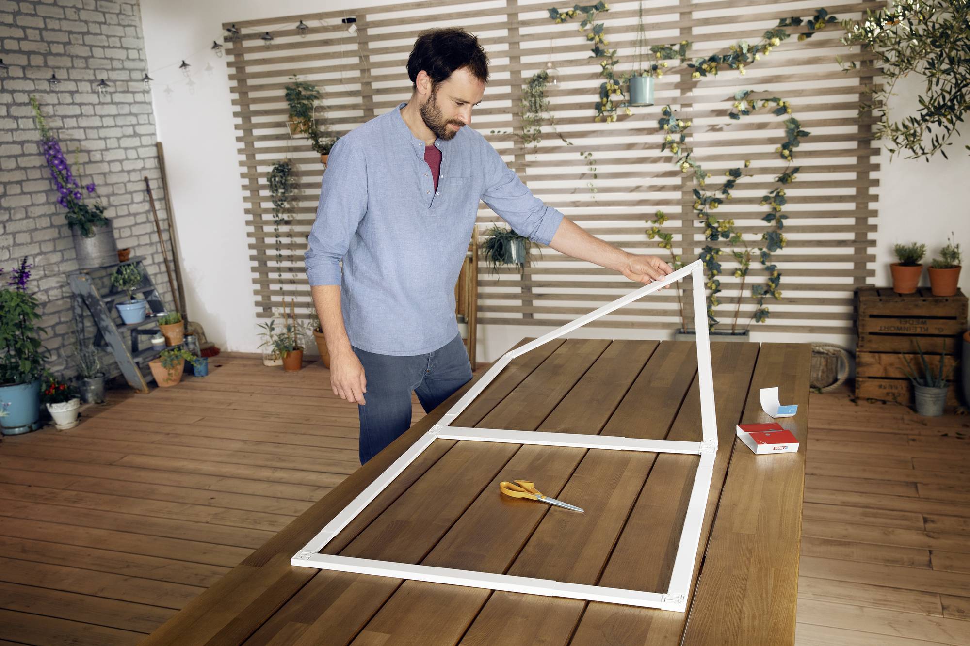 Een man in een blauw shirt staat op een terras en bouwt een houten frame. Planten zijn zichtbaar op de achtergrond.