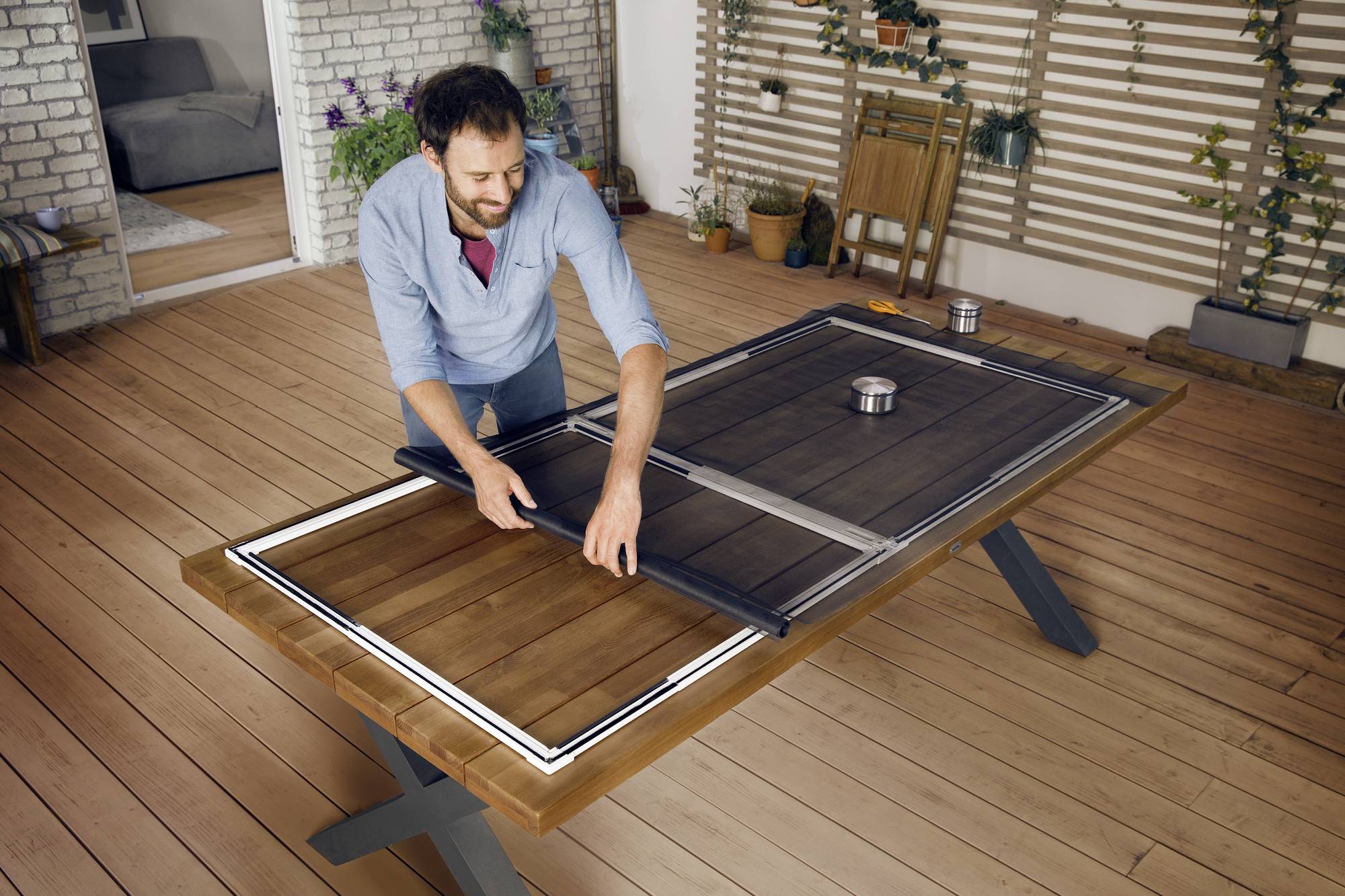 Een man monteert een tafelblad op een terras. Hij plaatst een donker paneel in het midden van een houten tafel.