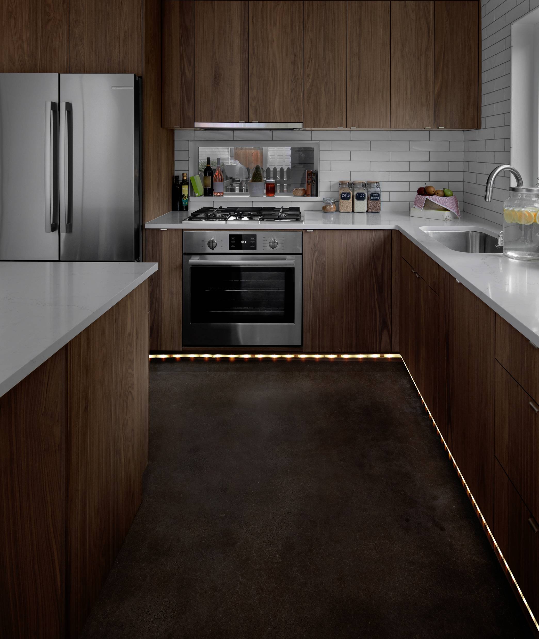 Moderne keuken met houten kasten, wit werkblad, roestvrijstalen apparaten en verlichting aan de vloer. Links staat een koelkast naast een fornuis.