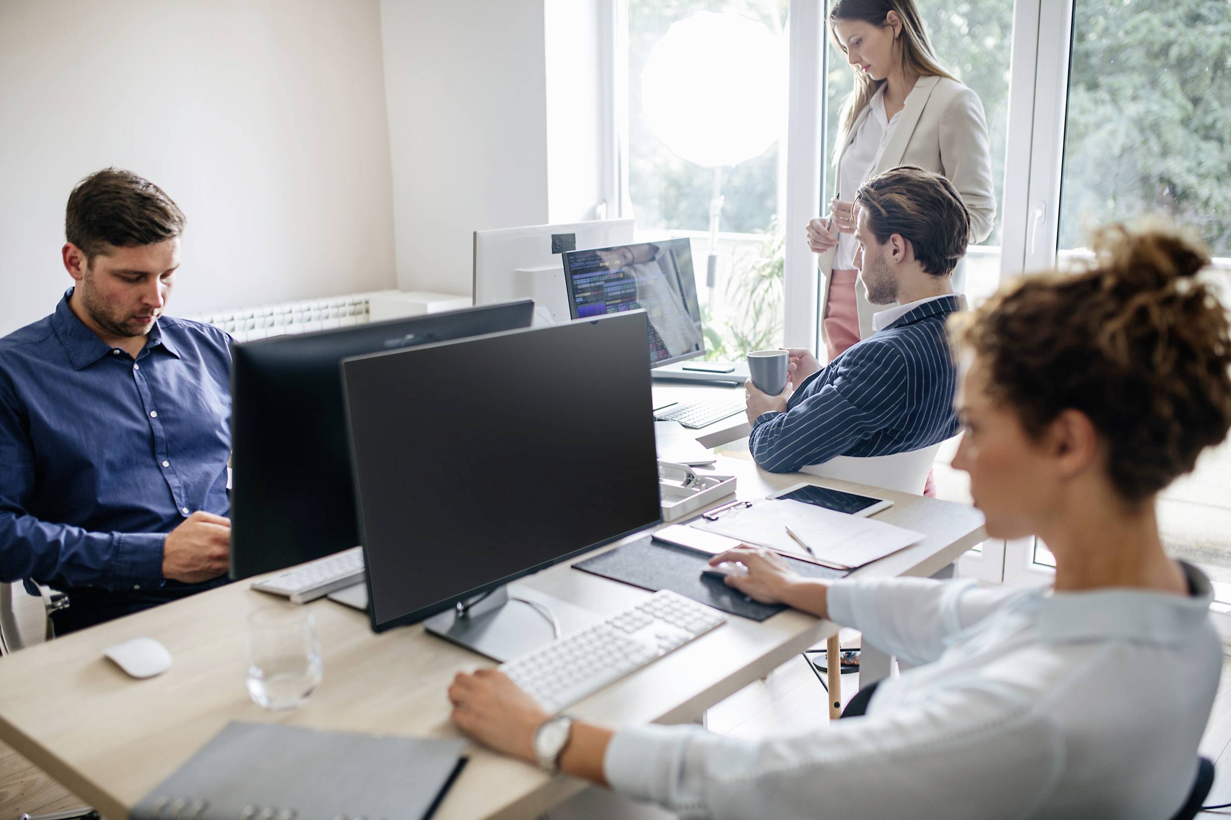 Vier personen werken op computers in een kantoor. Twee mannen zitten aan bureaus, een vrouw staat te praten en een andere vrouw is aan het typen.