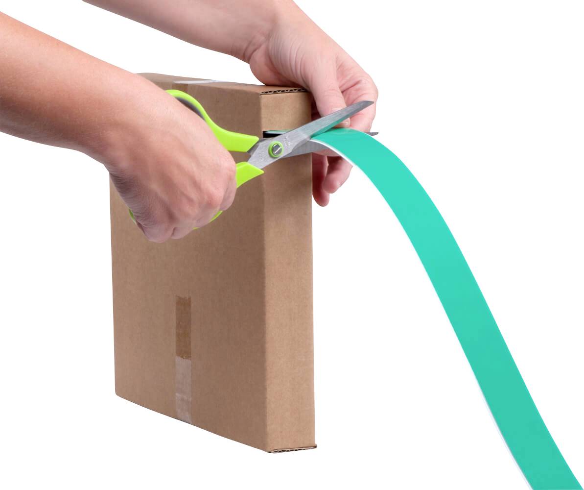 Een persoon knipt met een groene schaar een turquoise lint door op een bruine kartonnen doos.