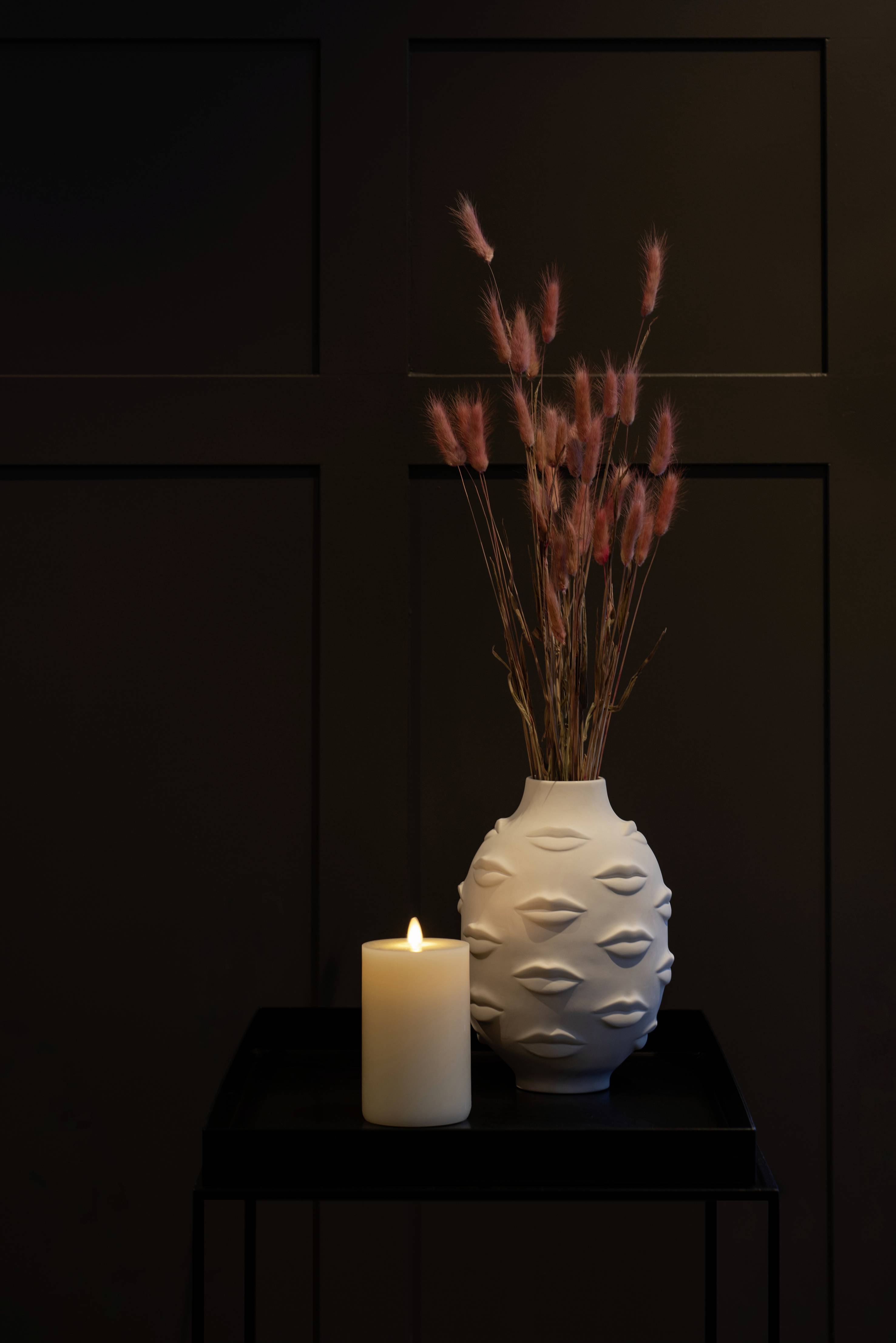 Een witte vaas met verhoogde patronen, naast een brandende kaars, staat op een zwarte tafel voor een donkere muur.