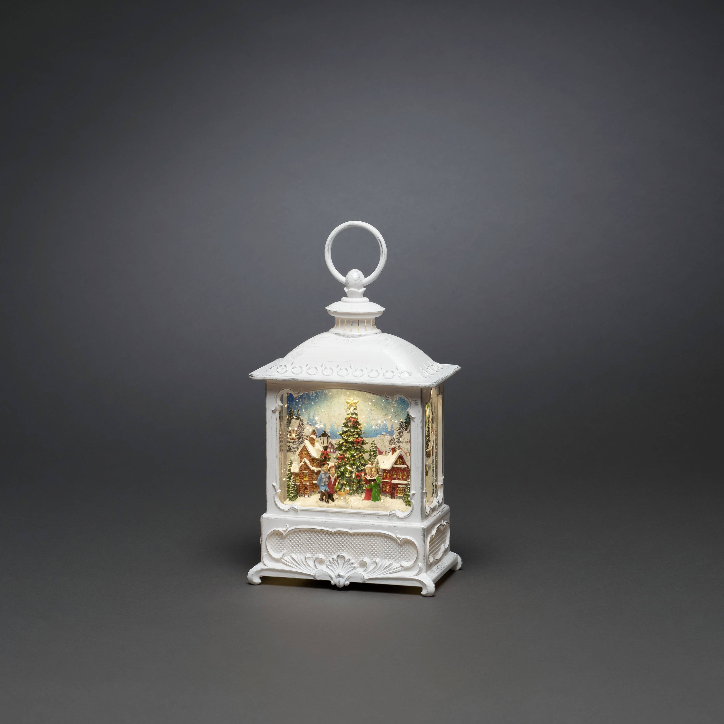 Een witte, decoratieve lantaarn met een kerstlandschap van binnen, toont een versierde boom en een feestelijk huis.