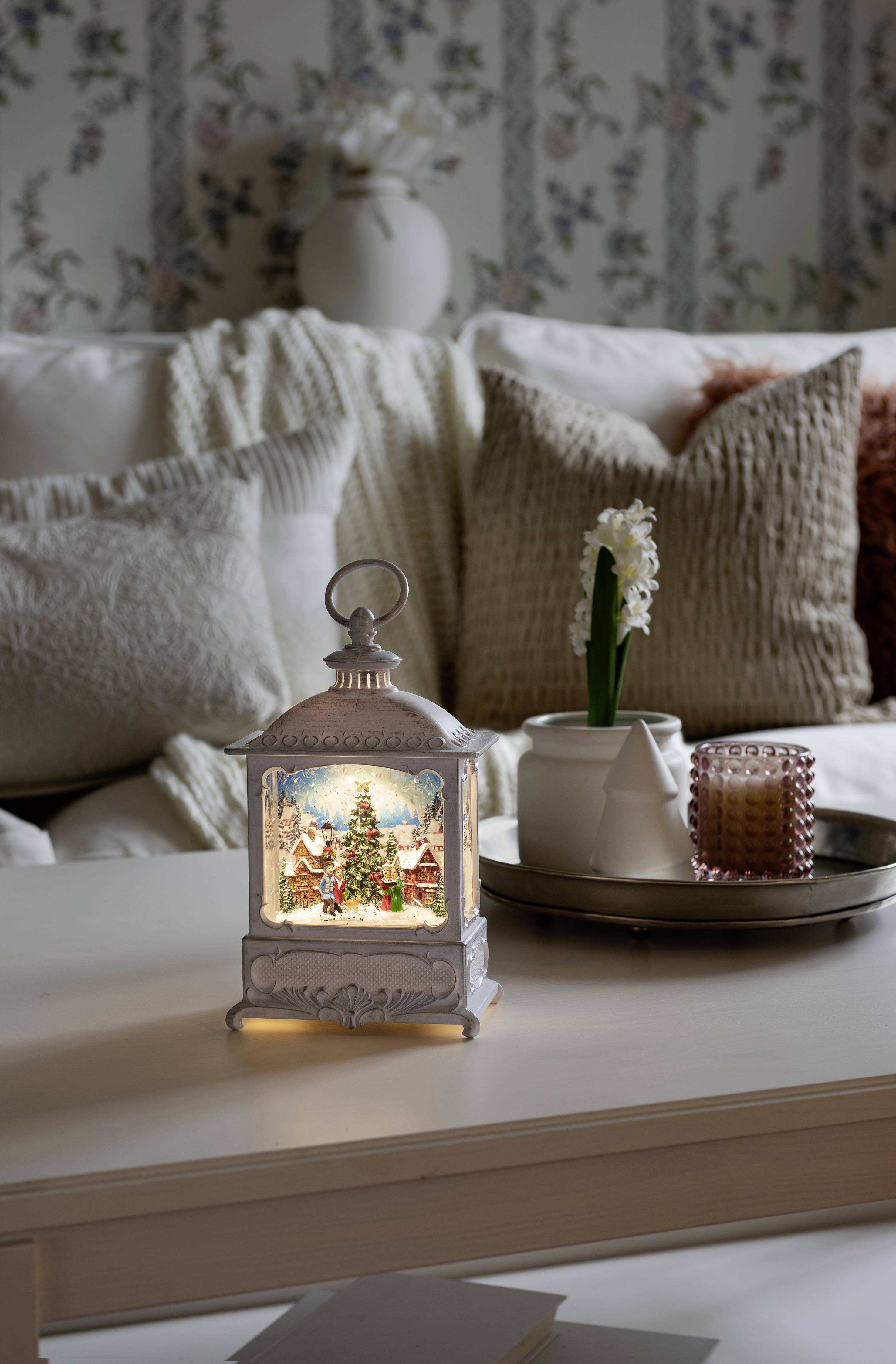 Lantaarn met sneeuwscène, verlicht op een tafel in de gezellige woonkamer. Achtergrond: decoratieve kussens en bloemenvaas.