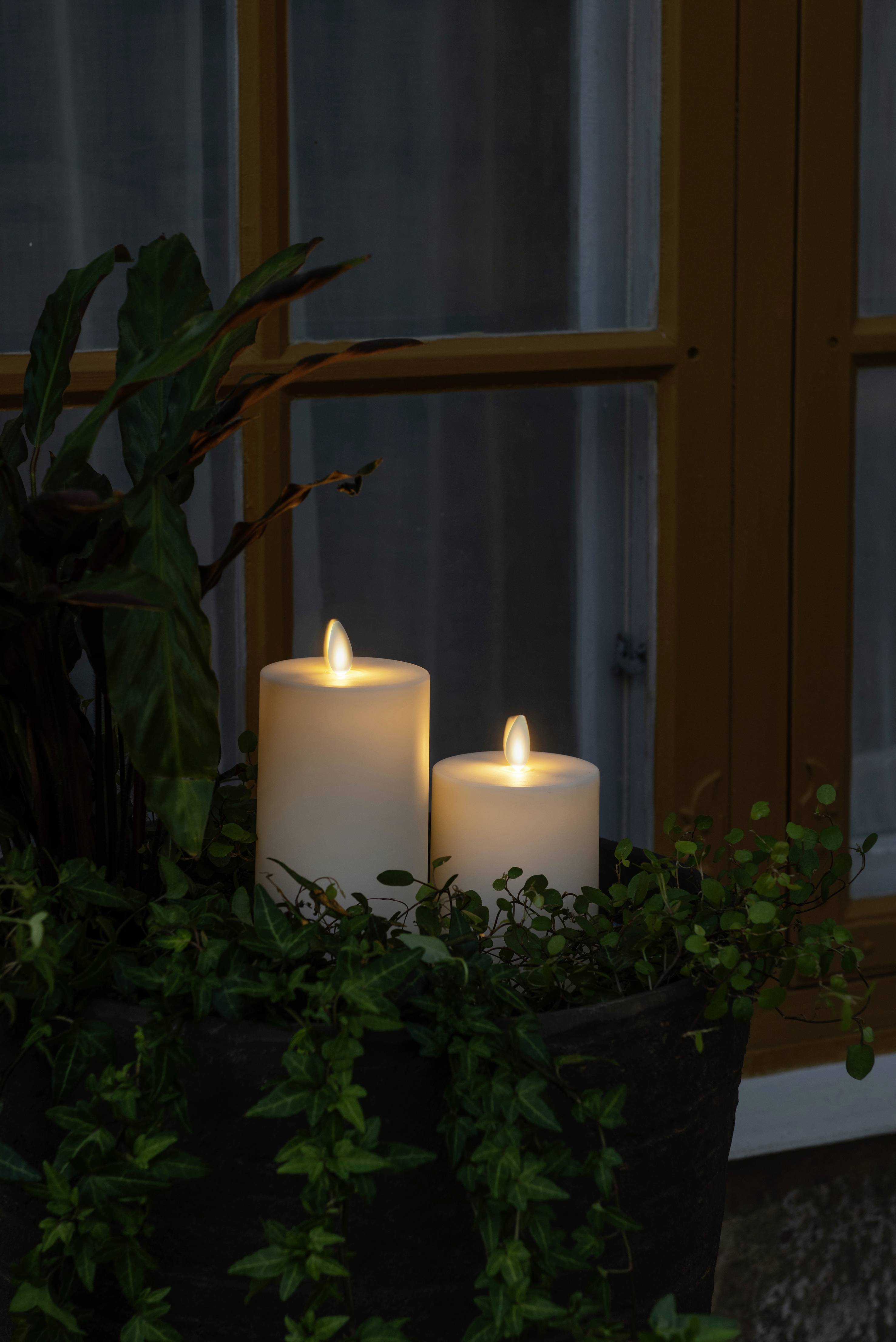 Twee brandende kaarsen staan naast een plant voor een raam. De kaarsen dompelen de scène in warm licht.