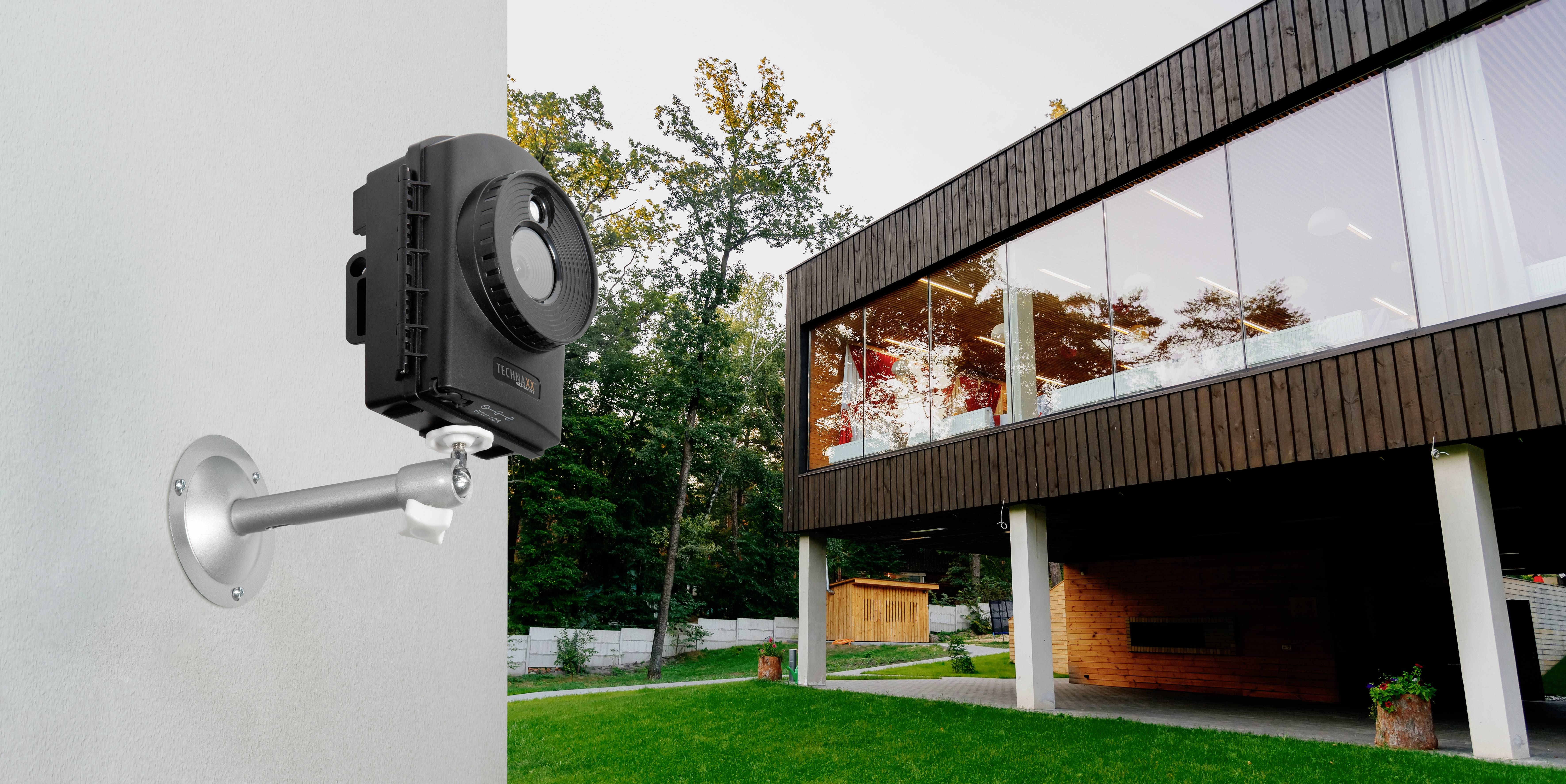 Een bewakingscamera is bevestigd aan de buitenmuur van een modern huis met een glazen gevel en houten elementen. Op de achtergrond bomen.