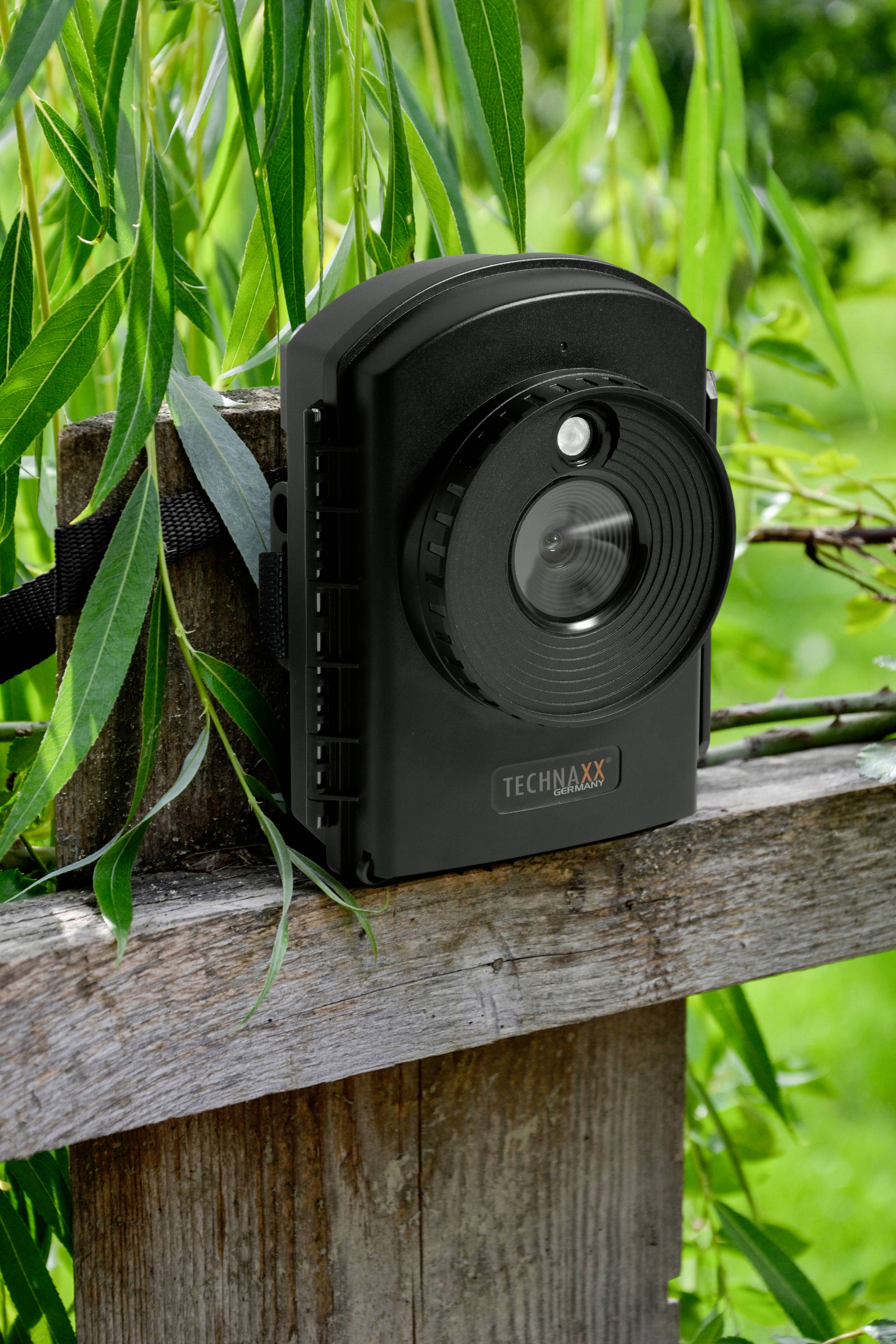 Een zwarte wildcamera is bevestigd aan een houten hek, omringd door groene planten. Op de camera staat 'TECHNAXX'.