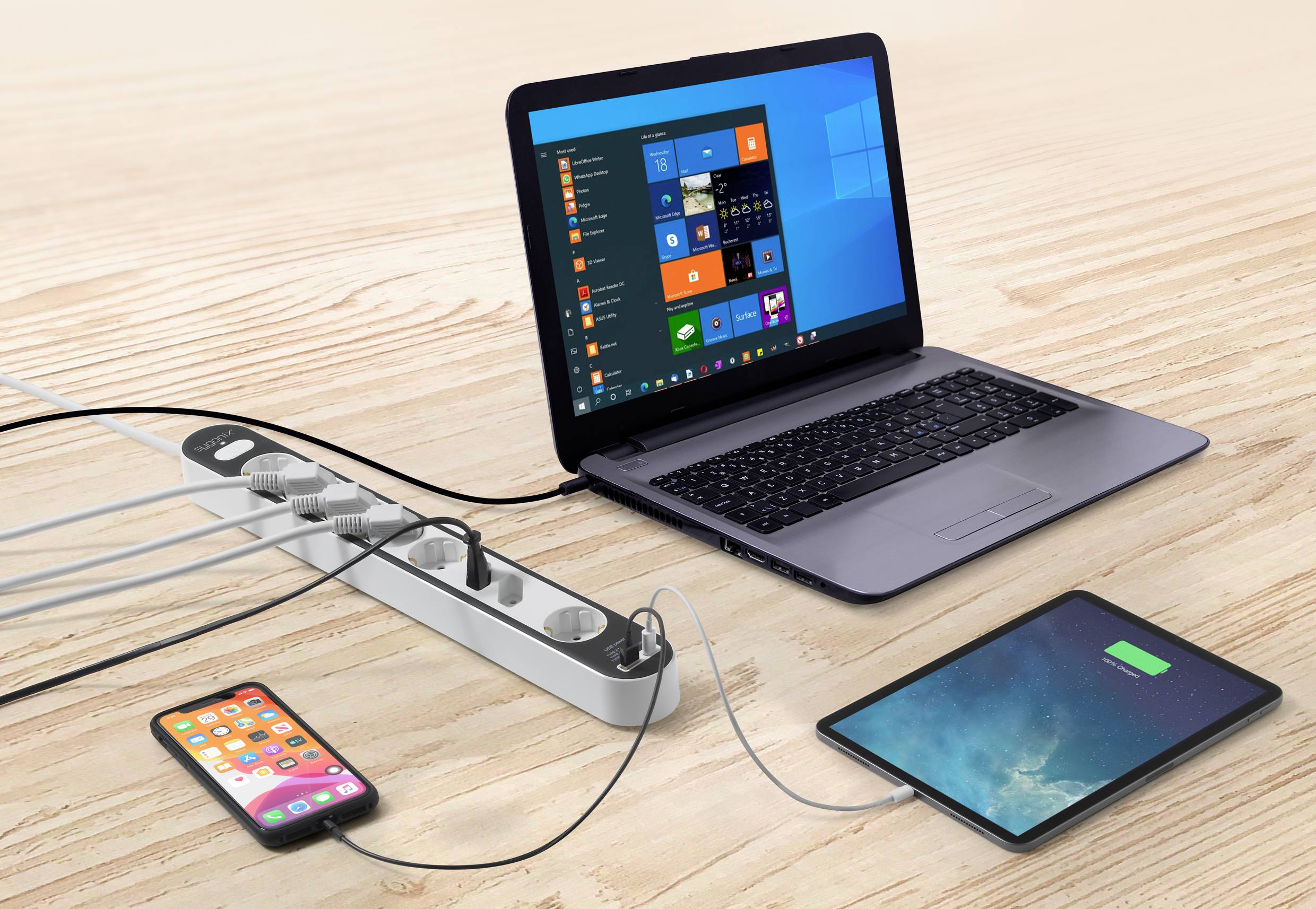Een laptop, een smartphone en een tablet zijn aangesloten op een stekkerdoos die op een houten tafel ligt.