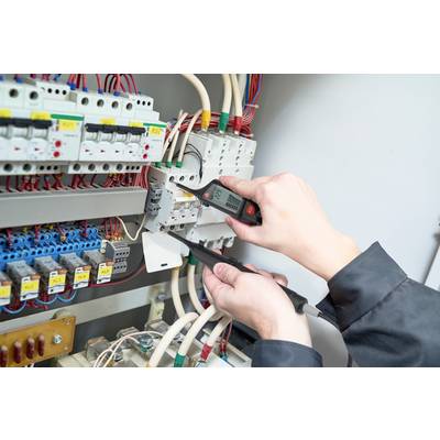 Persoon controleert elektrische schakeling in verdeelkast met multimeter. Hand houdt meetpennen tegen kabel. Veiligheidsuitrusting is zichtbaar.