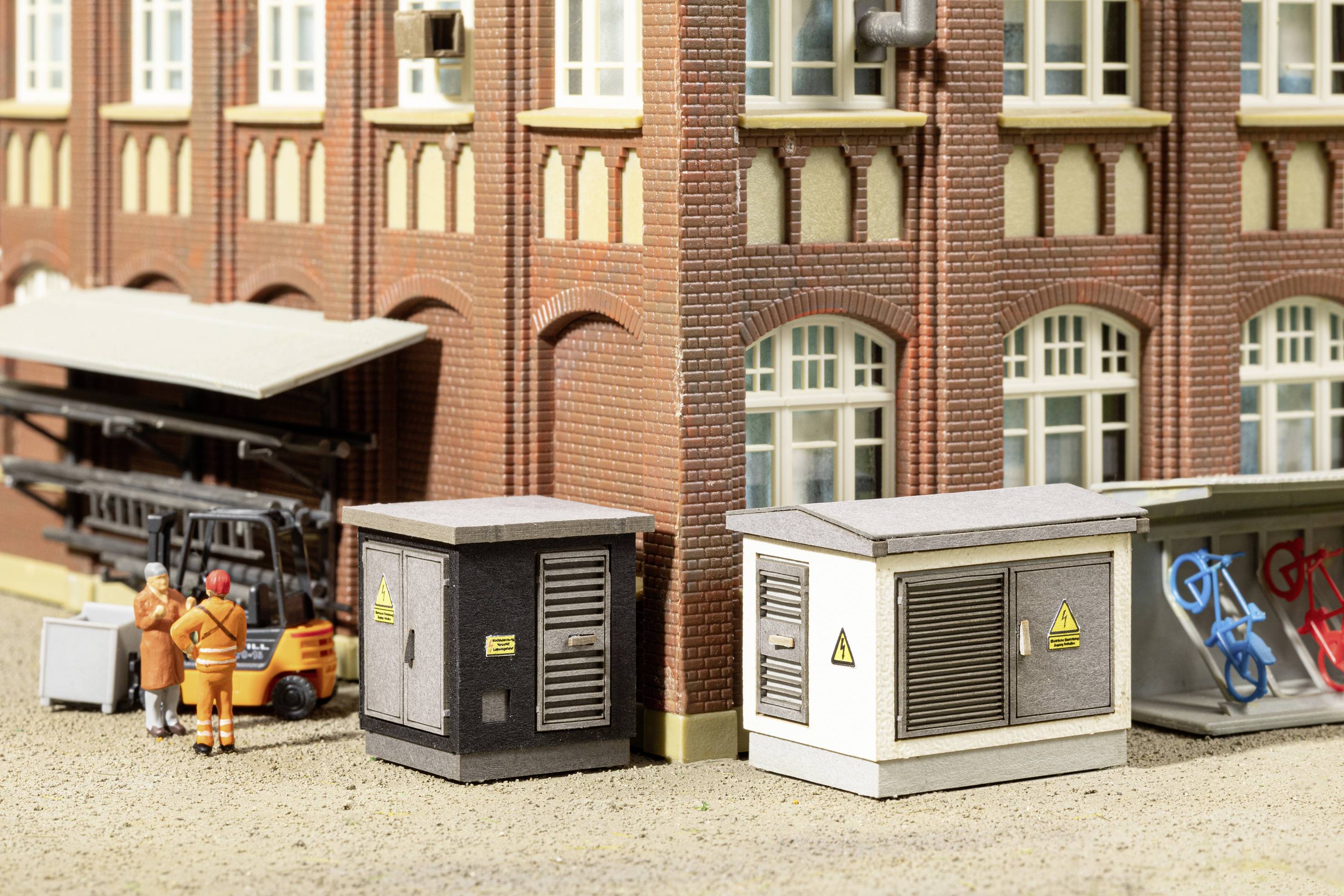Miniatuurscène met arbeiders naast een heftruck voor een gebouw. Twee kleine schakelkasten staan op de grond.