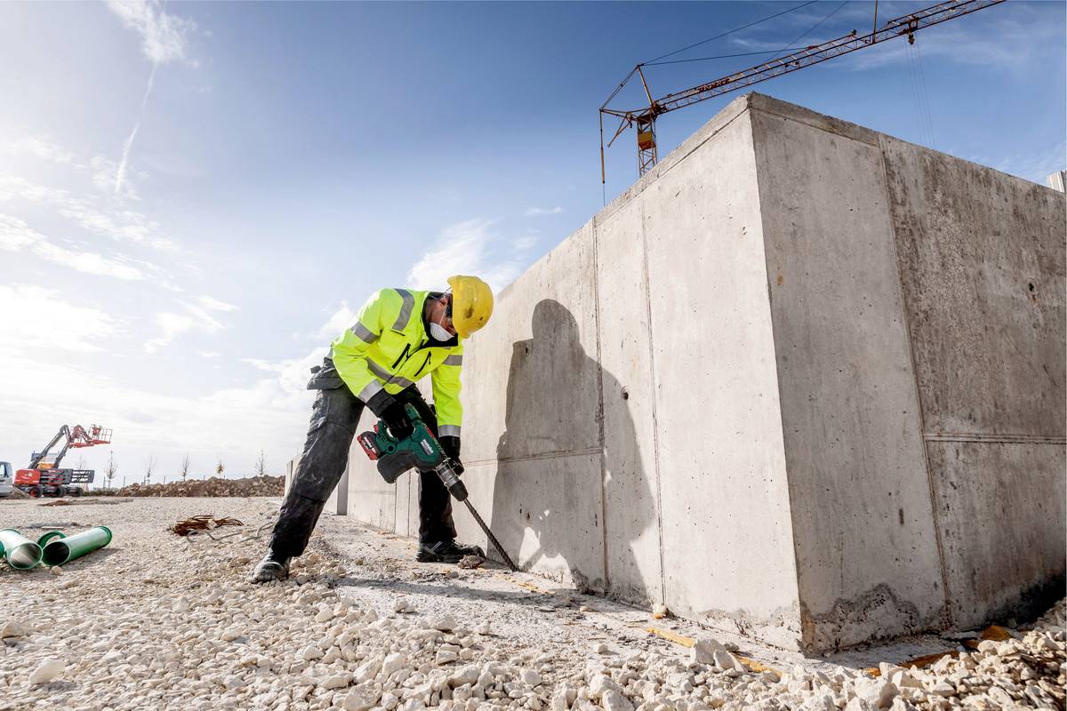 Bouwvakker in gele veiligheidskleding gebruikt een boormachine om een betonblok te bewerken op een bouwplaats met een kraan.