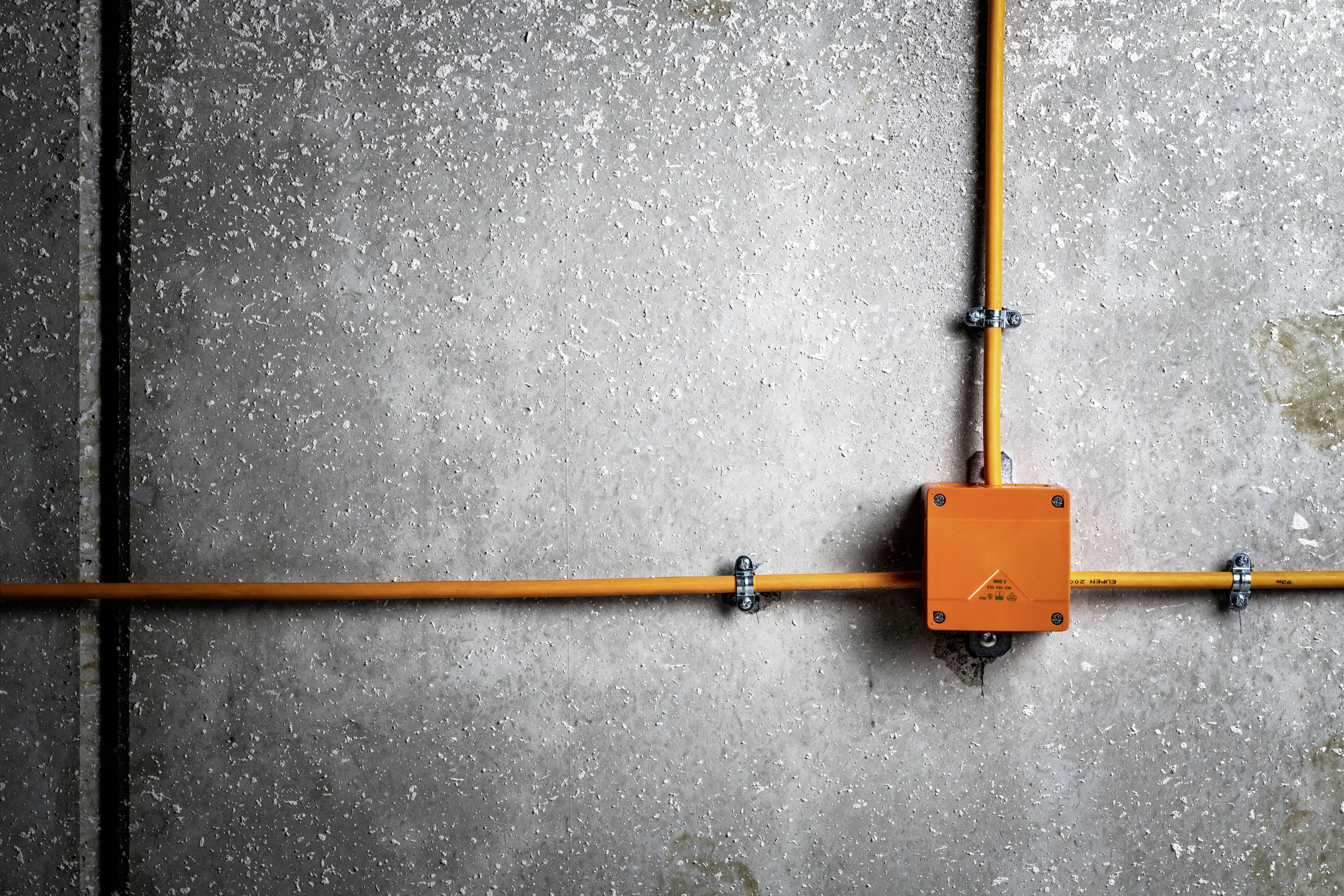 Oranje kabels en dozen langs een grijze betonnen muur, die elkaar in een rechte hoek kruisen. Toont een gestructureerd leidingsnetwerk op een industriële oppervlakte.