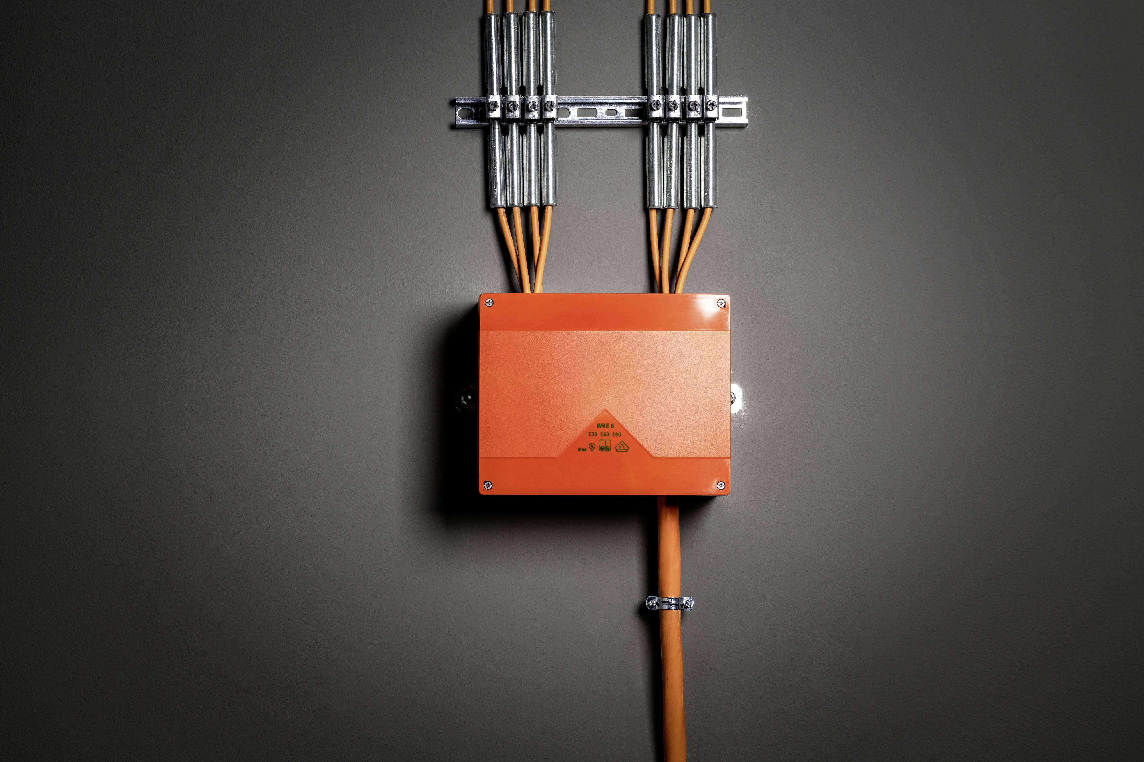Een oranje verdeelkast met meerdere kabels loopt langs een grijze muur omhoog. De kabels zijn netjes gerangschikt.