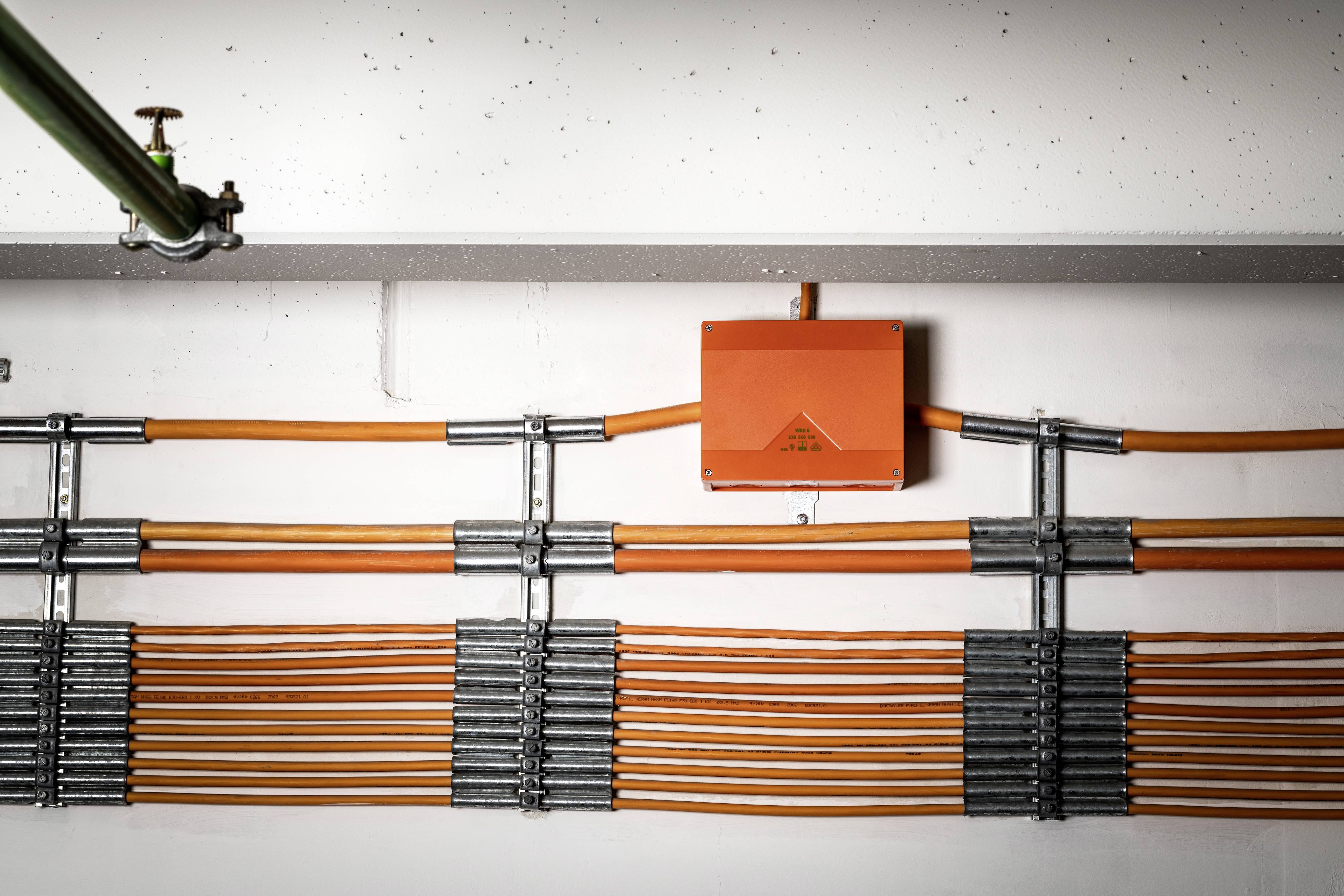 Oranje kabelgoten lopen horizontaal langs een witte muur, verbonden door zilveren metalen beugels.