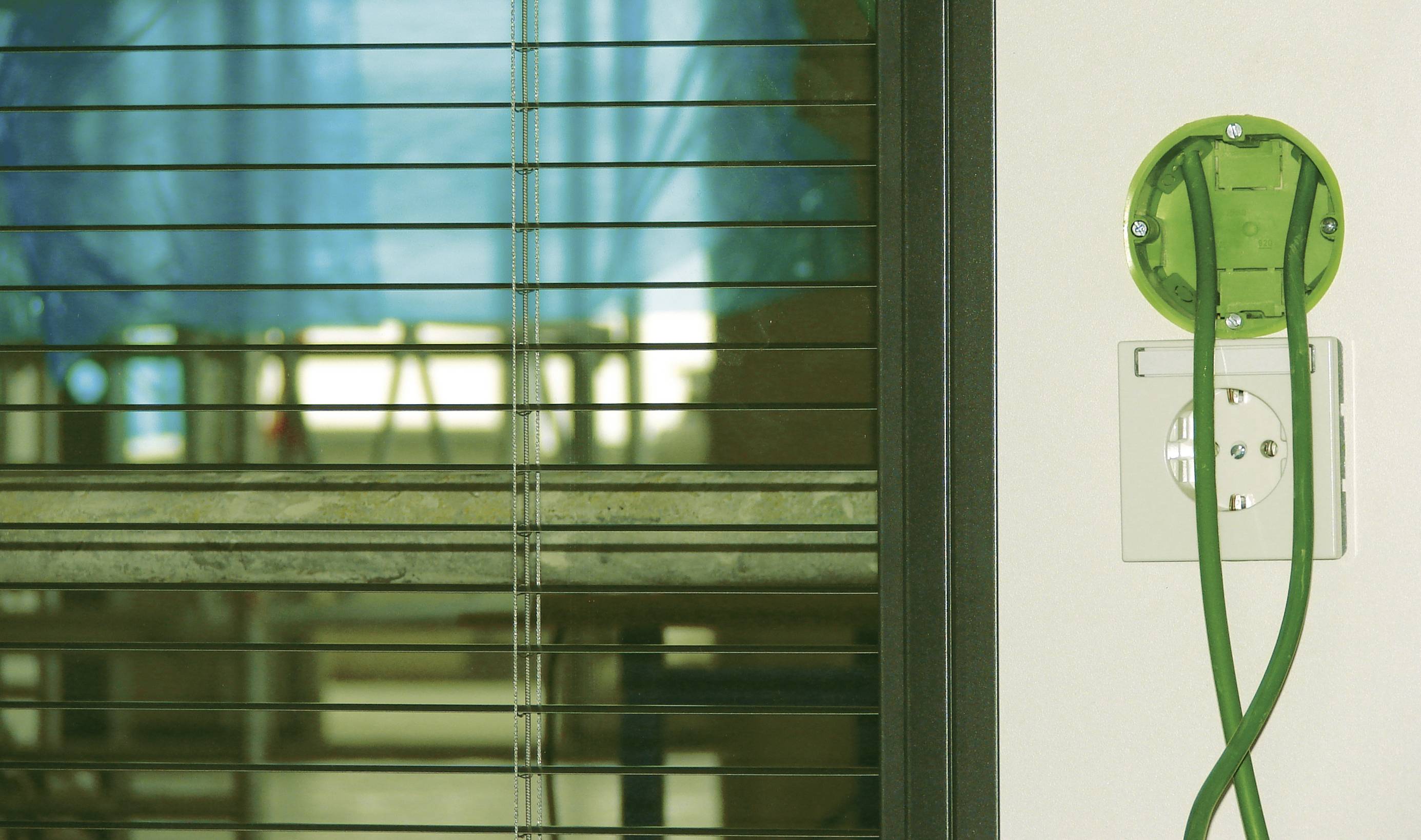 Een groene stroomstekker hangt aan een wit stopcontact naast een raam met halfgesloten jaloezieën, op de achtergrond een steiger.