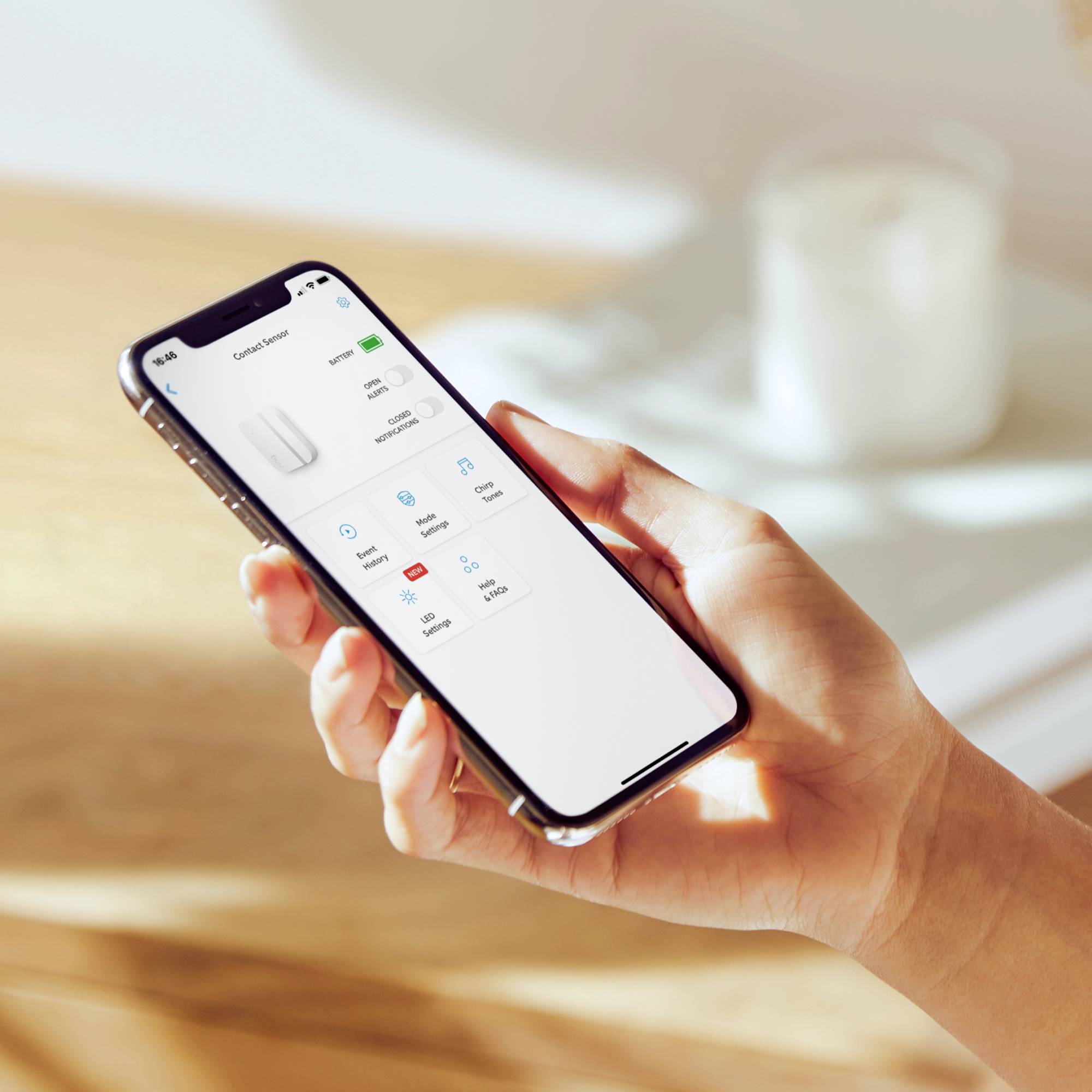 Een hand houdt een smartphone vast waarop een app met opties voor een elektronisch apparaat wordt weergegeven. Op de achtergrond een onscherpe tafel.