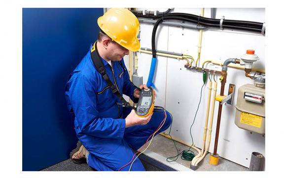 Een technicus in blauwe werkkleding en een gele helm gebruikt een meetapparaat om buizen en leidingen in een technische ruimte te inspecteren.