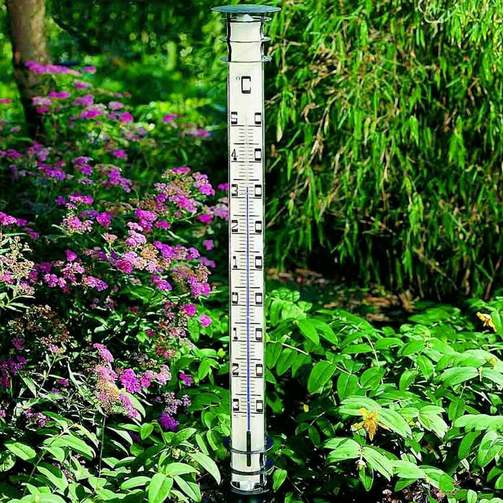 Een thermometer toont 25°C in een tuin met roze bloeiende planten en groen gebladerte op de achtergrond.