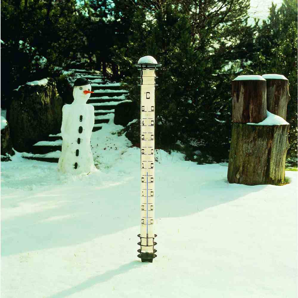 Een sneeuwpop naast een hoge meetlat in de sneeuw geeft een hoogte van 40 cm aan. Op de achtergrond zijn bomen en houten balken te zien.