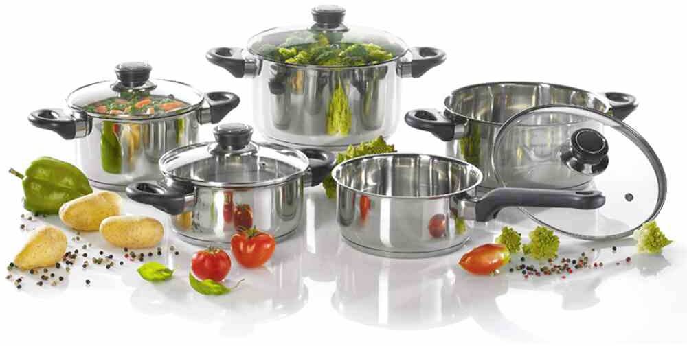 Een set van vijf glanzende roestvrijstalen potten en pannen, gevuld met groenten zoals broccoli en tomaten, op een reflecterende oppervlakte.