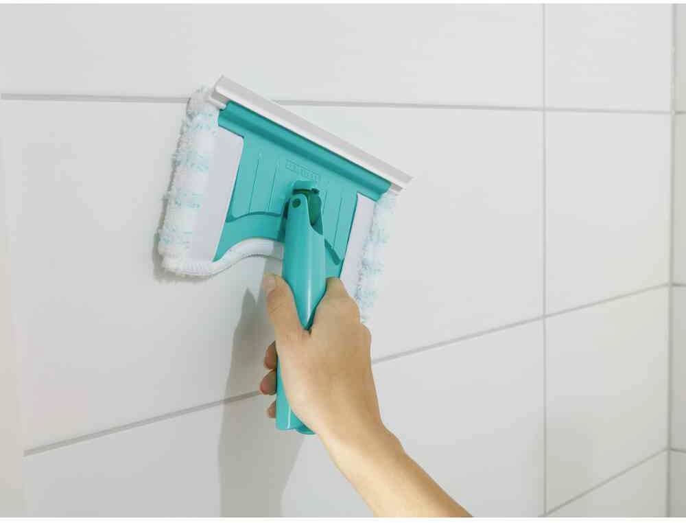 'Hand houdt raamwisser met blauwe handgreep vast aan witte betegelde muur. Wisser reinigt de muur.'