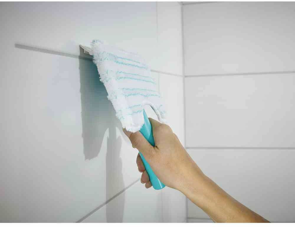 Een persoon reinigt witte tegels met een dweil. De dweil heeft een blauwe steel en een afveegbare witte oppervlakte met blauwe strepen.