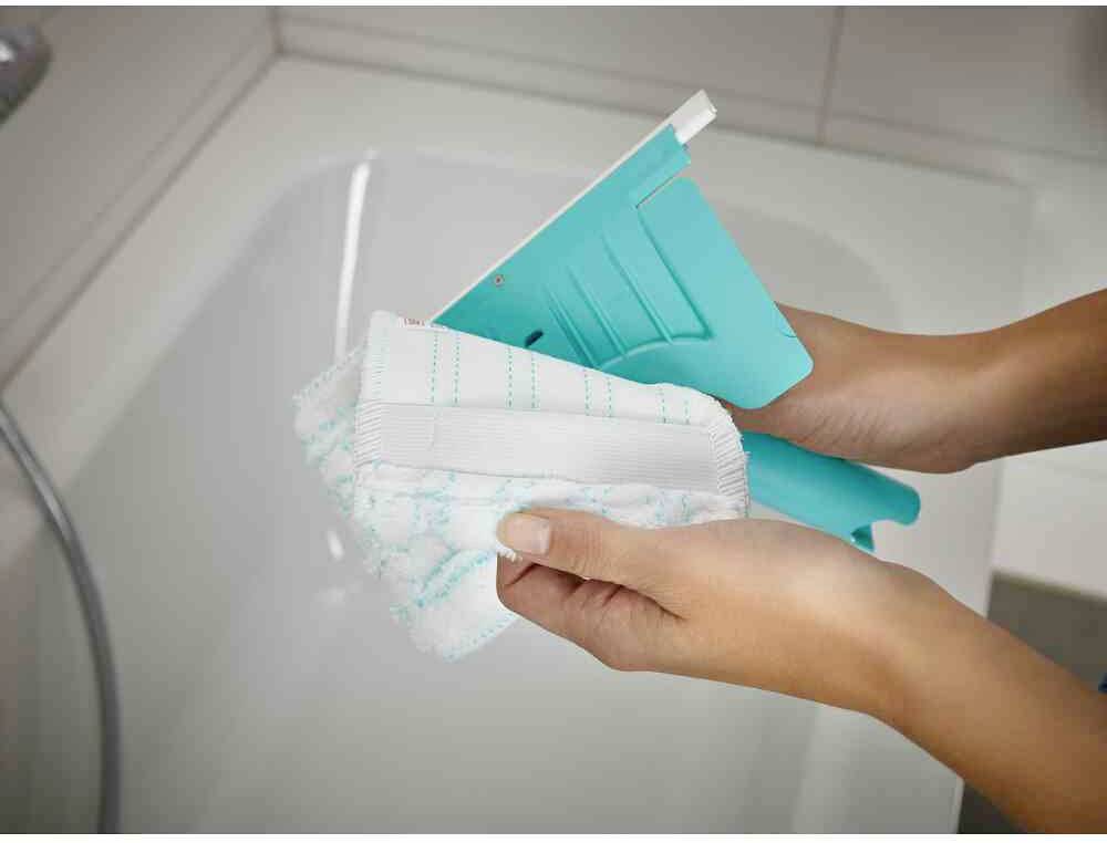 Een persoon houdt een turquoise tegelreiniger vast met een wit reinigingspad voor een witte badkuip.