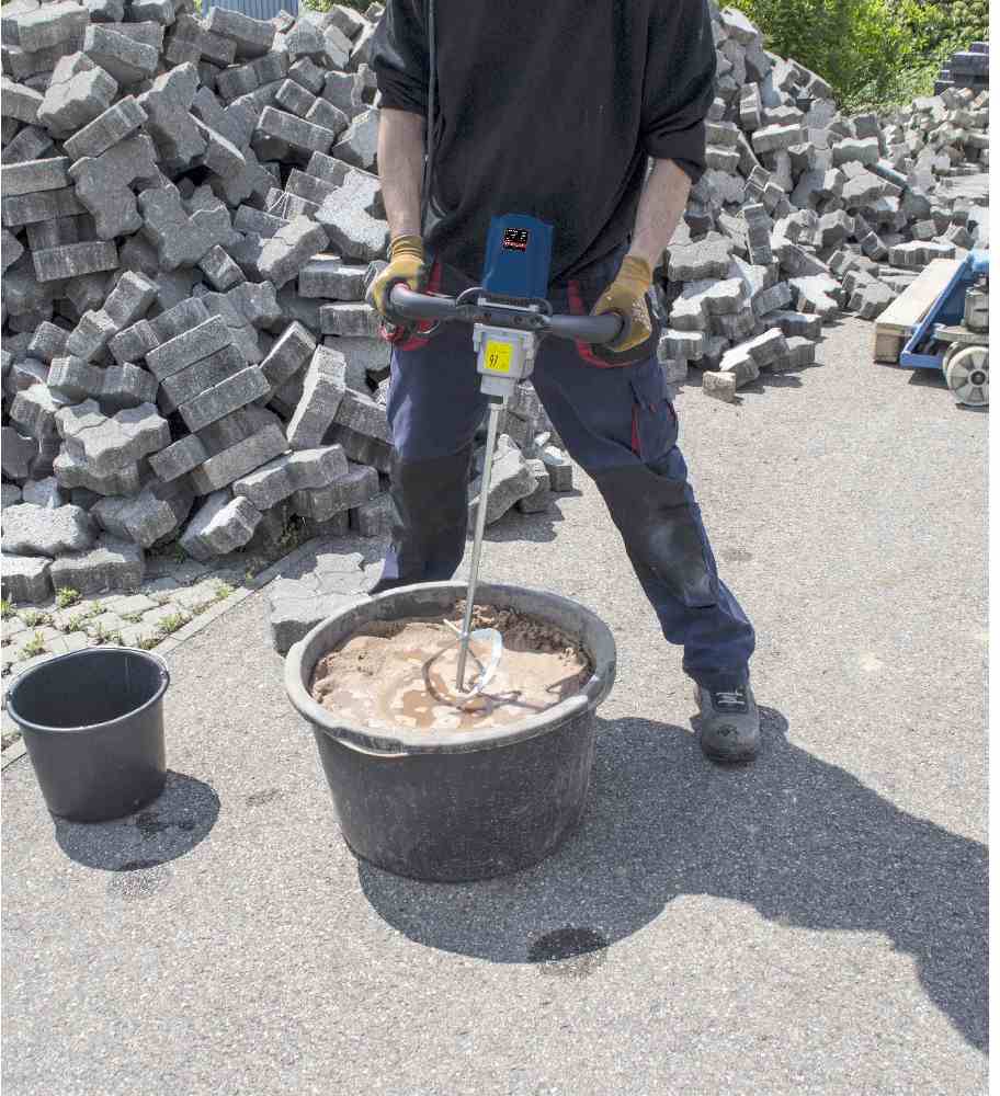 Een persoon mengt beton in een emmer met een elektrisch roerapparaat, omringd door gestapelde bestratingstegels op een bouwplaats.