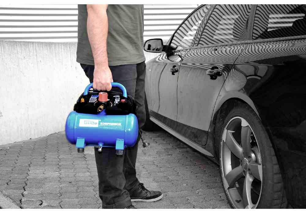 Een persoon draagt een kleine, draagbare blauwe compressor naast een geparkeerde auto op een bestrate ondergrond.