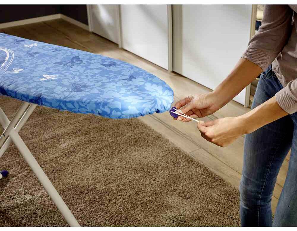 Een persoon trekt aan een blauwe strijkplankhoes met een bloemenmotief, die staat op een bruine vloerbedekking in een kamer.