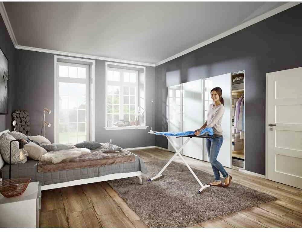 Een persoon strijkt een overhemd op een strijkplank in een modern ingericht slaapkamer met grijze muren en een houten vloer.