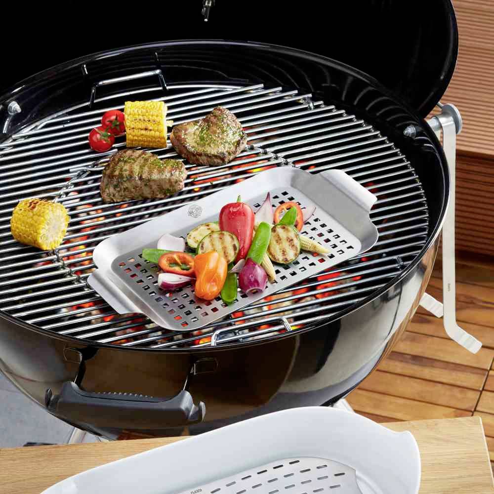 Een ronde houtskoolbarbecue met gegrilde groenten, maïskolven en stukken vlees. De barbecue staat op een houten terras.